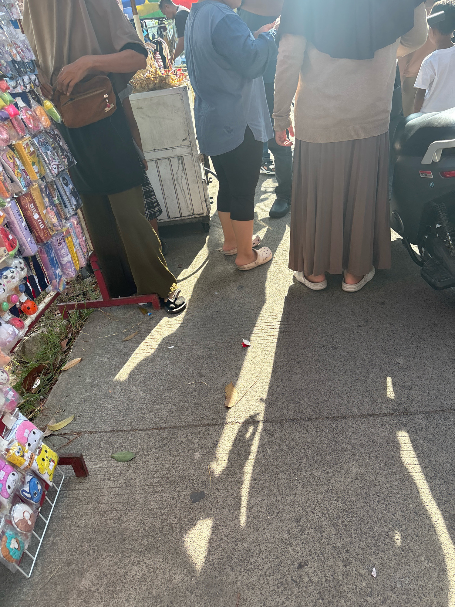 The image captures a street scene in Telagamurni, Indonesia, on a sunny day. In the foreground, a person is standing near a display of various merchandise, which appears to be small toys or accessories packaged in clear plastic. The items on display are colorful and varied. To the right of the display, a person wearing a tan sweater and a long brown skirt is standing next to a parked motorcycle. Further left, another person in a blue long-sleeved shirt and black pants is visible. A third person, wearing a tan shirt and dark pants with a small brown bag slung across their body, is partially visible on the far left. The ground is paved with concrete, and shadows from the people and overhead structures are cast across it. The overall scene suggests a bustling market or roadside stall environment.
