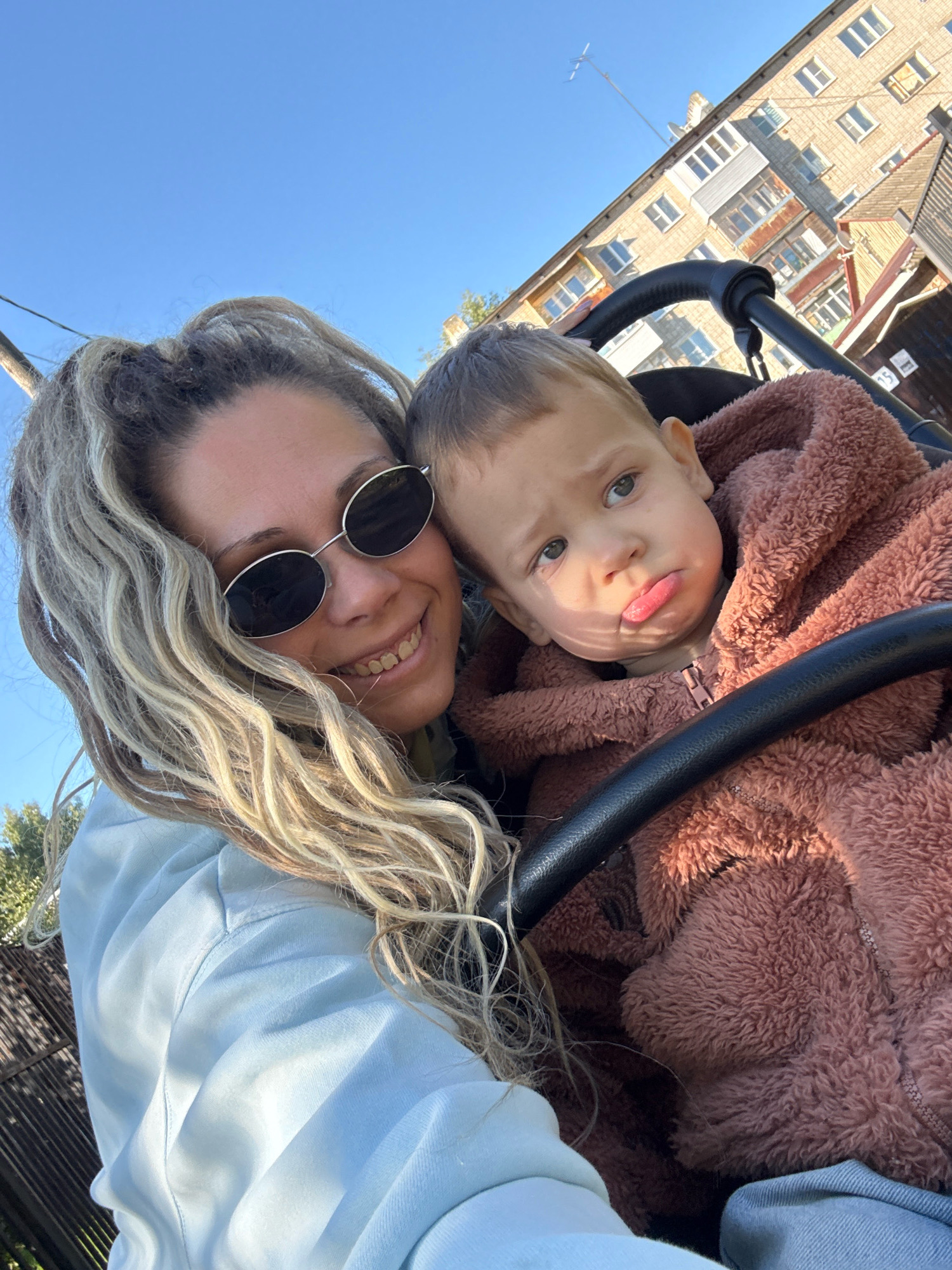 In a selfie taken outdoors in Rusanovo, Russia, a woman with blonde dreadlocks and sunglasses smiles at the camera while holding a young child in a stroller. The child, bundled in a pink fuzzy jacket, makes a pouty face. Behind them, a multi-story brick building with balconies and a wooden structure are visible against a clear blue sky.