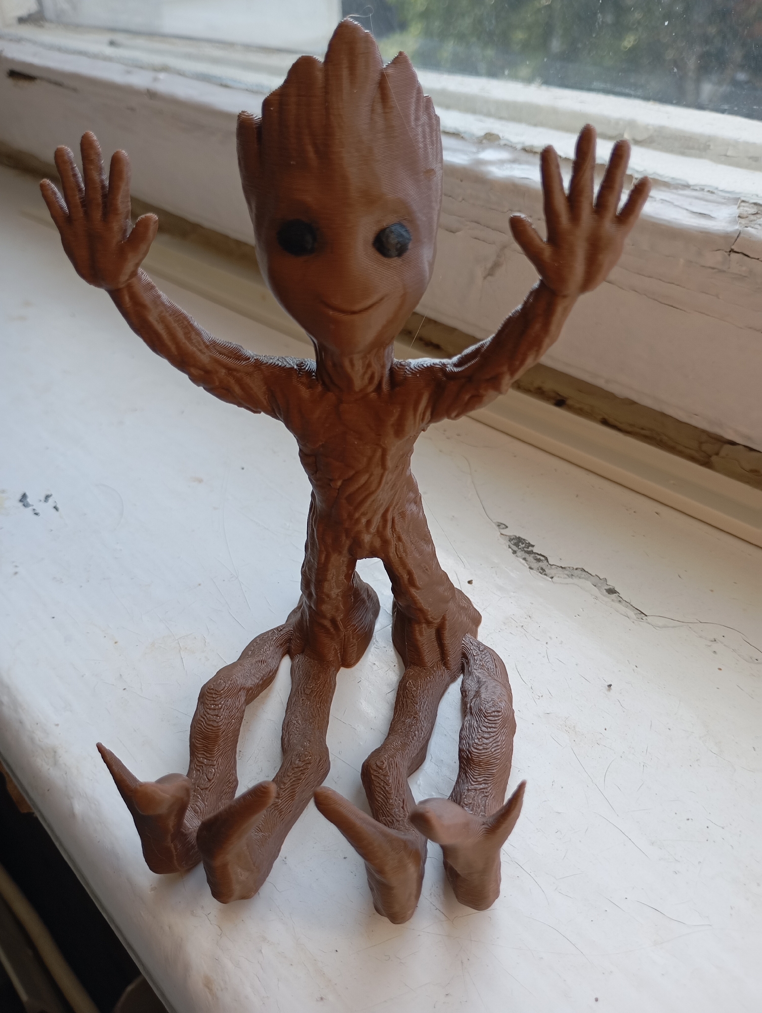 A brown, 3D-printed figurine of Groot, a character from Marvel comics, is sitting on a white windowsill. The figurine is positioned facing forward, with its arms raised and hands open, as if waving. Its body is textured to resemble bark, and it has black dot eyes and a smiling mouth. The windowsill is aged and shows signs of wear, with peeling paint and cracks. Behind the figurine, a window with a faded wooden frame is visible, suggesting an indoor setting. The background outside the window is blurred, with hints of greenery and trees, indicating it is likely daytime. There is no visible indication of the city of Gorki-2, Russia.