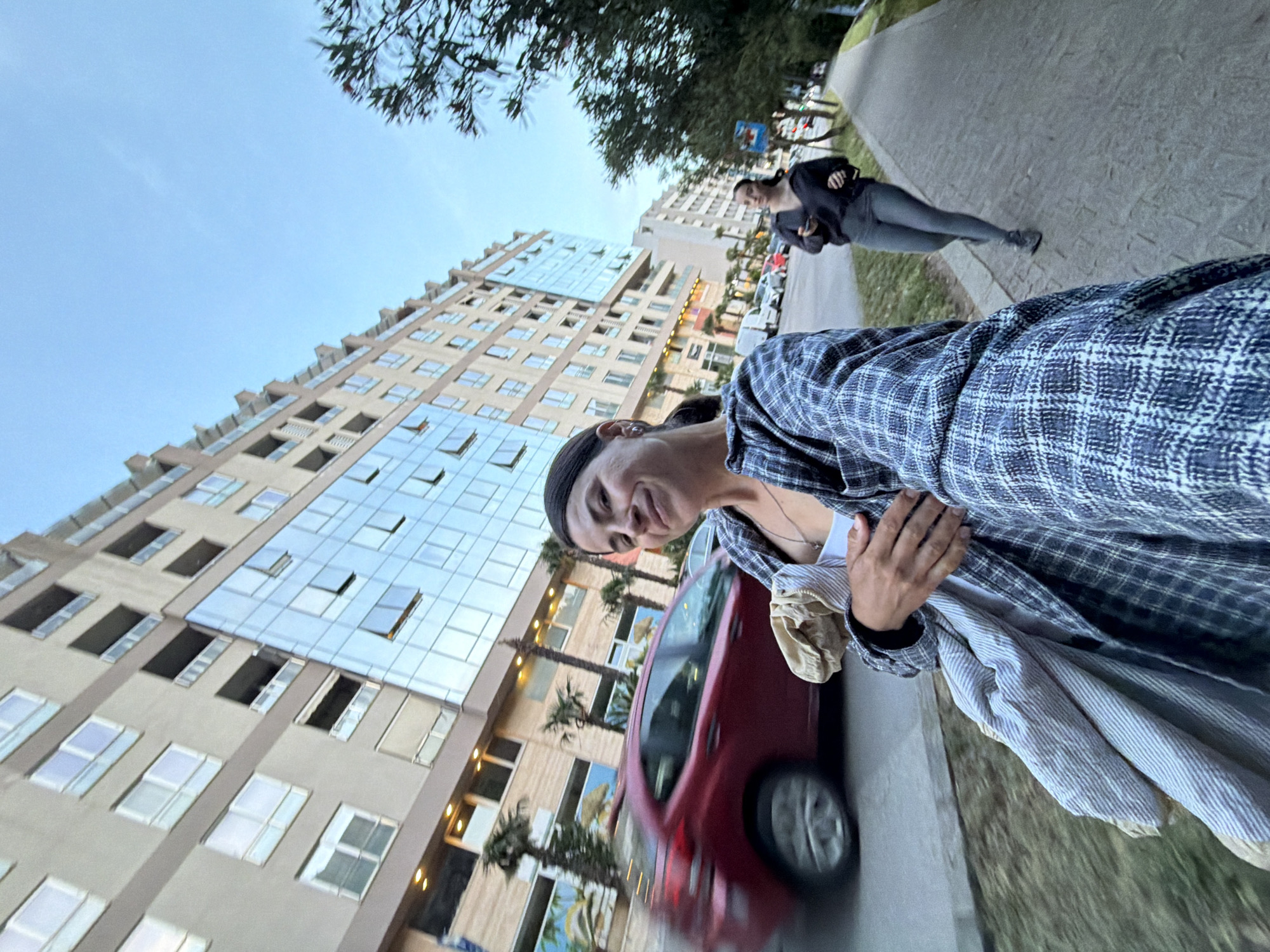 The image captures a street scene in Budva, Montenegro, taken from a low angle with a wide field of view, creating a slight distortion. The sky is clear and bright blue, suggesting good weather during late afternoon or early evening.
In the foreground and to the right, a person, likely female, looks towards the camera with a relaxed smile. She has dark hair and wears a blue and white plaid overshirt over a white top. Her left hand is visible, holding a striped piece of clothing. A red car is partially visible to her left, slightly blurred as if in motion or out of focus.
Further back on the sidewalk, another person, also likely female, walks away from the camera, wearing dark clothing. She is positioned near some trees and a partially obscured road sign.
The left side of the frame is dominated by a tall, modern building with many windows, some of which are open. The building's facade reflects the sky, and some ground-level windows show warm, artificial lighting. Palm trees are visible in front of the buildings, adding to the Mediterranean urban setting. A paved sidewalk runs along the right side of the image, separated from the street by a curb and some grass. No discernible text is visible on signs or buildings.
