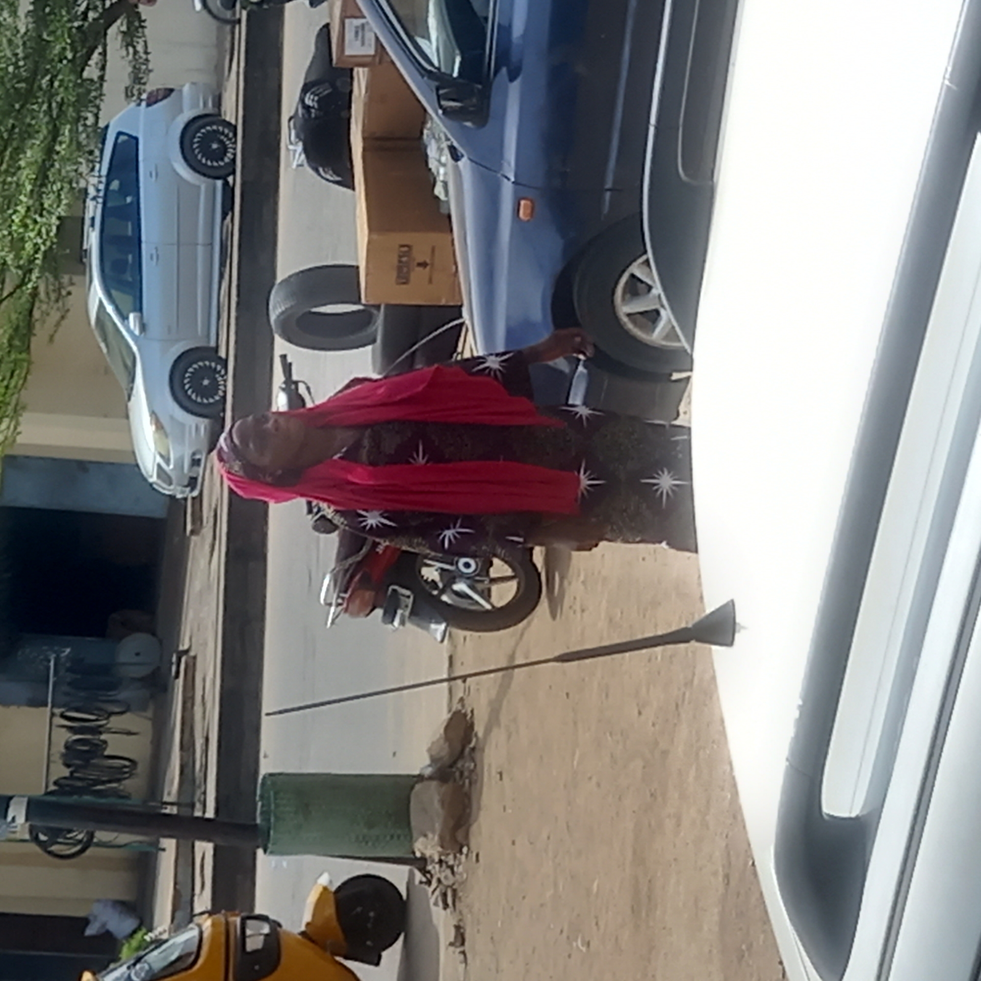 The image shows a street scene in Dadi Riba, Nigeria. An older woman wearing a red head covering and a dark, star-patterned dress stands near a blue vehicle and a motorcycle. There is a white car partially visible in the foreground. Behind the blue vehicle are tires and a cardboard box. A white car is parked further down the street. There is a green post and a yellow vehicle on the left side of the image. The scene is in daylight.