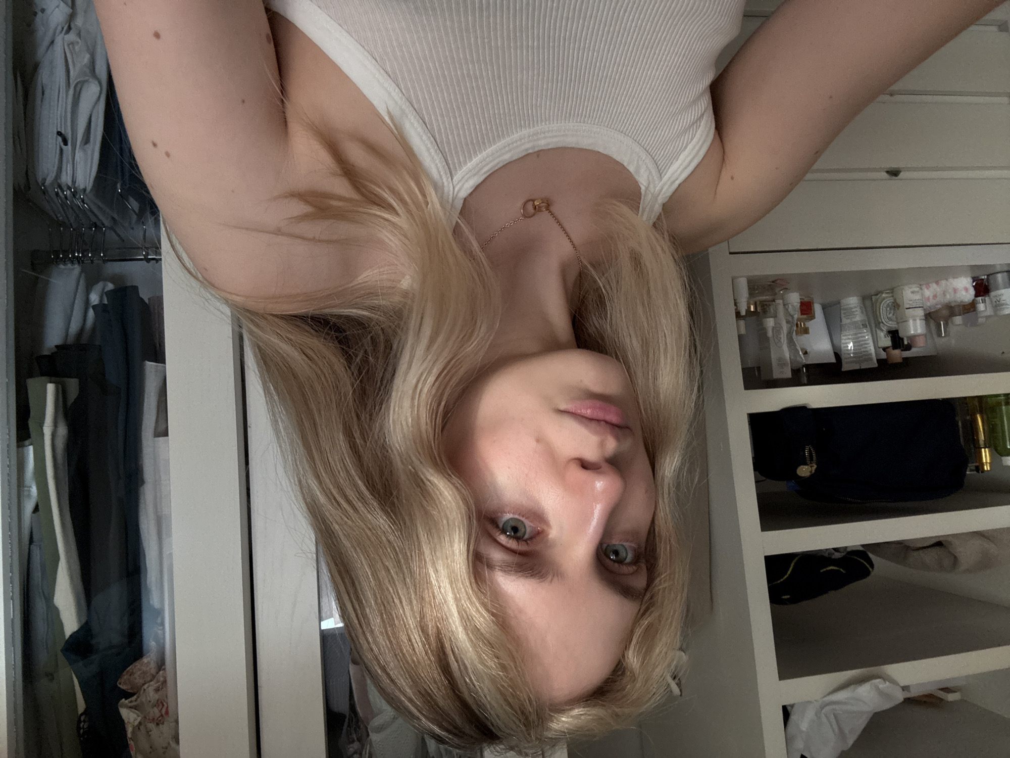 The image shows a young woman with blonde hair, pale skin, and green eyes, wearing a white ribbed tank top and a delicate gold necklace. She is positioned upside down in the frame. In the background, there is a white wardrobe. The left side of the wardrobe contains hanging clothes in shades of gray and dark green. The right side features shelves with toiletries, perfumes, and bags. The setting appears to be indoors. There is no indication of the location being in Mexico City or any other location.