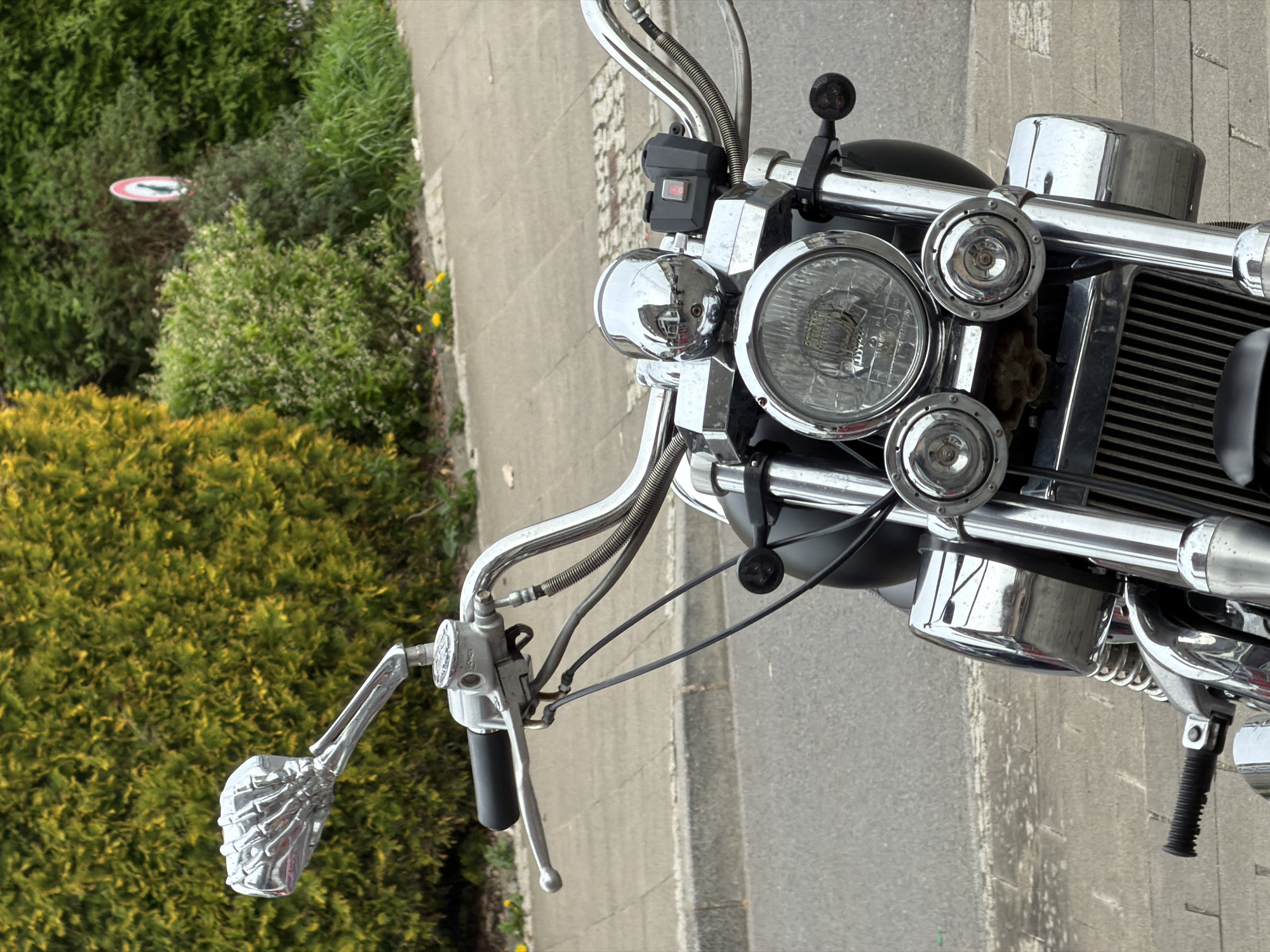 The image shows the front of a motorcycle parked on a paved road, likely a sidewalk or parking area adjacent to a road. The motorcycle has a chrome finish on many of its parts, including the handlebars, headlight housing, and engine components. The left handlebar features a distinctive chrome skeleton hand grip. The background includes a variety of green foliage and bushes. A red and white sign is visible in the background, suggesting the location is in Esch-sur-Alzette, Luxembourg. The environment appears to be a residential or suburban area.