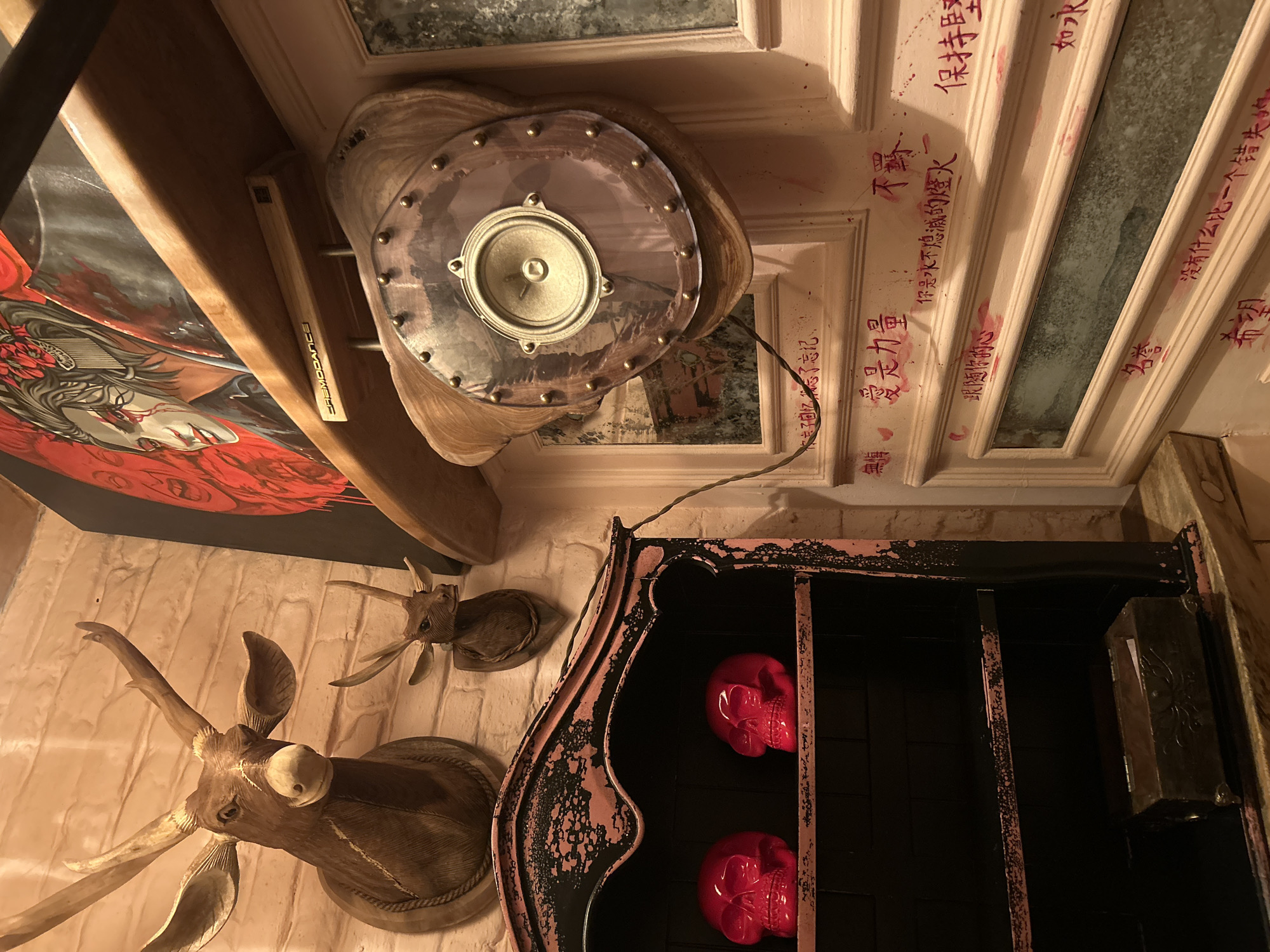 A low-angle shot shows a cluttered interior. On the wall, there are two mounted deer heads. A cabinet with two red skull-shaped objects inside is adjacent to the deer heads. Above this, a speaker is mounted on a decorative panel. A large piece of art is partially visible to the left. The ceiling is decorated with red handwritten characters. No location is possible from the image.