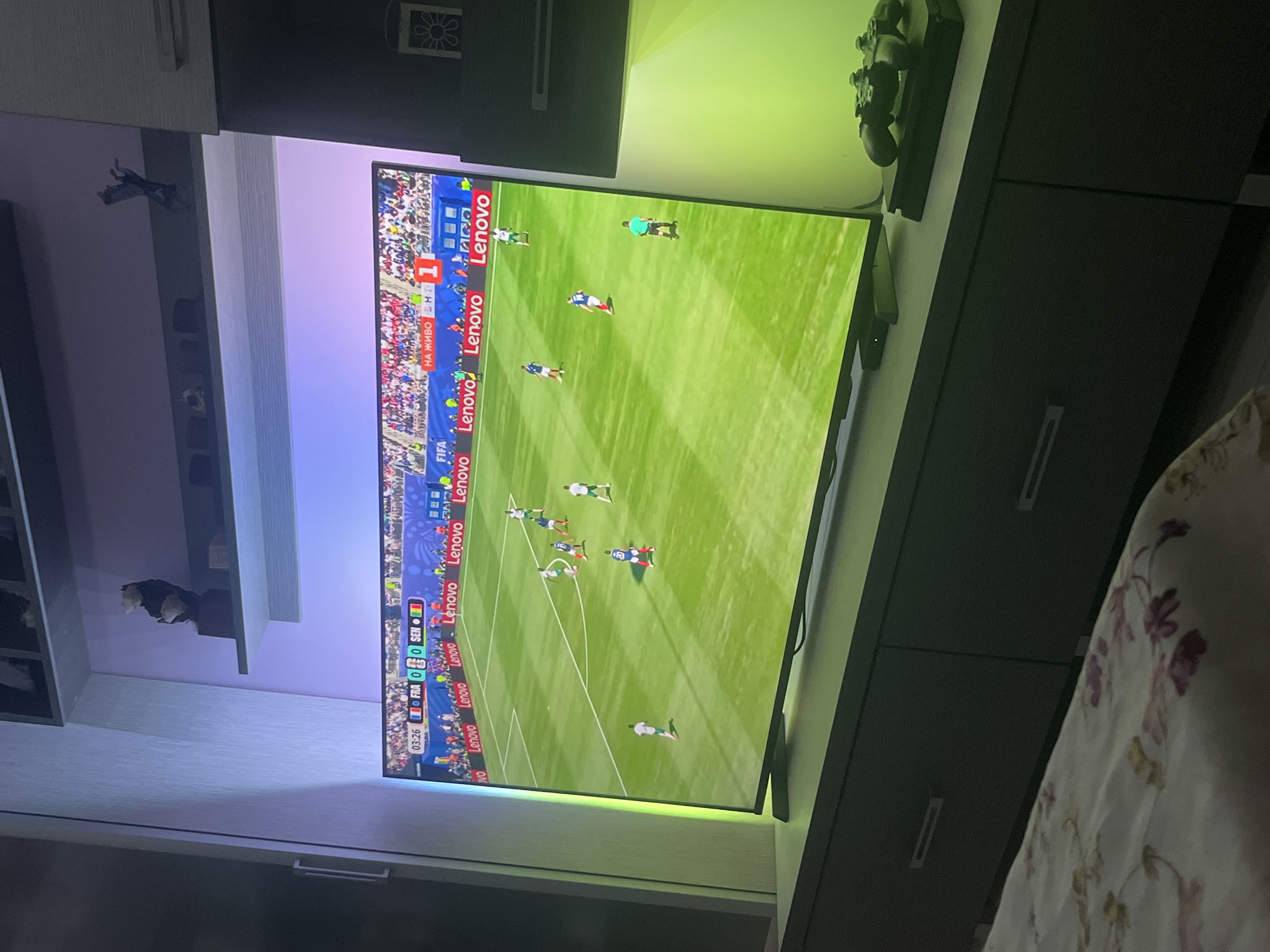 The image captures a television screen displaying a soccer match. The score is tied at 0-0 between France (FRA) and Senegal (SEN) at 03:26 minutes into the game. Several players in white and blue uniforms are visible on the green field. A large "Lenovo" advertisement runs along the bottom of the screen. The television is placed on a dark gray cabinet, which has two drawers. To the right of the television, on the cabinet, rests a black video game controller. The overall setting appears to be a living room or bedroom, with a bed in the foreground covered in floral patterned bedding. The background shows shelves with decorative items, and ambient colored lighting emanating from behind the television and on the wall. There is no discernible location in Burgas, Bulgaria.