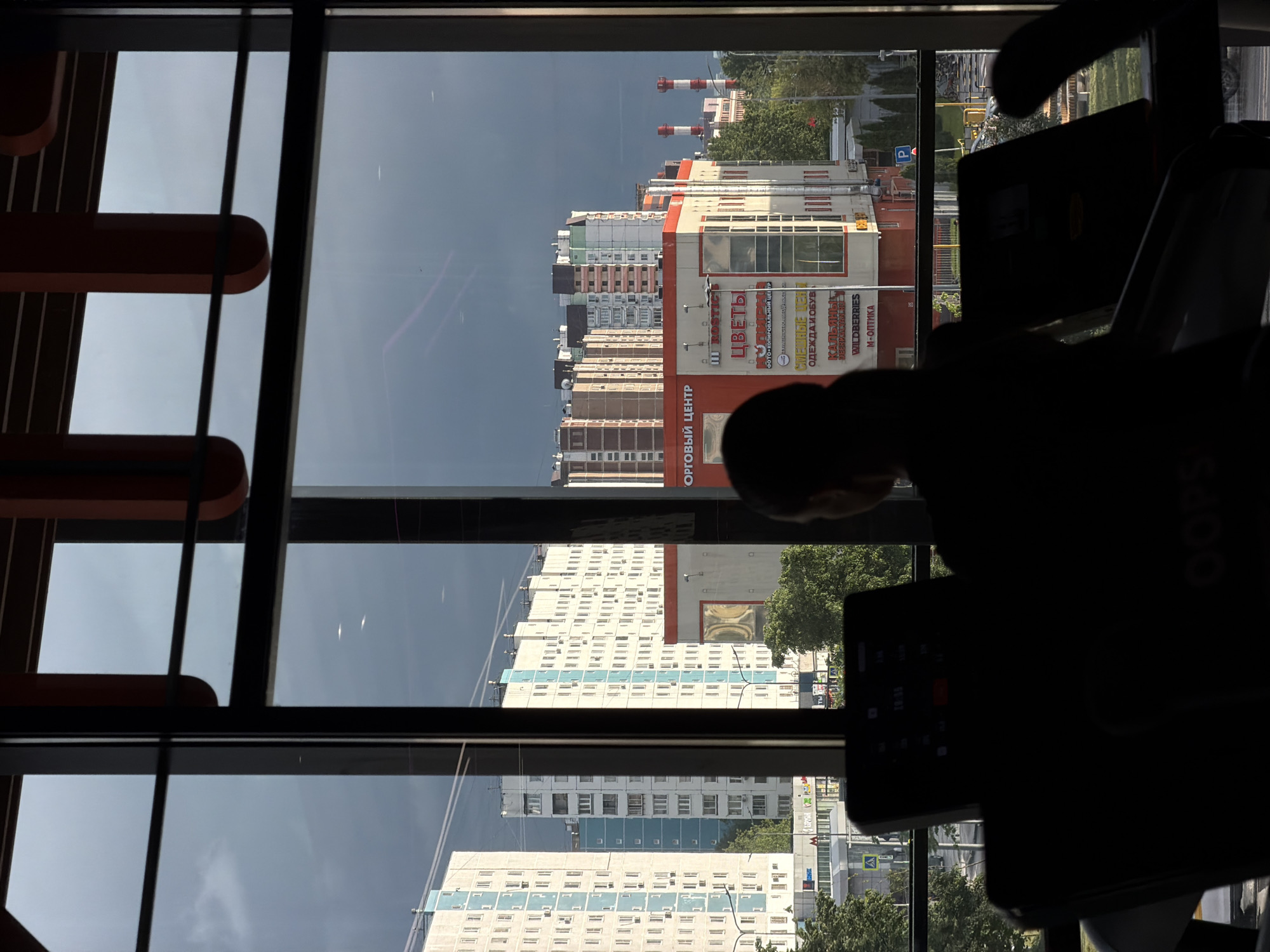 A person on a treadmill is seen through a multi-paned window in what appears to be a fitness center. The window looks out onto a city view, with several tall buildings visible. One of the buildings has a large sign in Russian that reads "ОРГОВЫЙ ЦЕНТР," which translates to "Wholesale Center". Other smaller signs on the same building mention "ЦВЕТЫ" (flowers), "ФОТО-КОПИРОВАЛЬНЫЙ ЦЕЇ ТР" (photo-copying center), "СМЕШНЫЕ" (funny), "ОДЕЖДАИ ОБУВ" (clothing and shoes), "КАЛЬЯНЫ" (hookahs), "WILDBERRIES," and "М-ОПТИКА" (M-Optics). In the background, there are industrial chimneys with red and white stripes. The sky is overcast. Based on the architectural style and the signs in Russian, the location is Moscow, Russia.