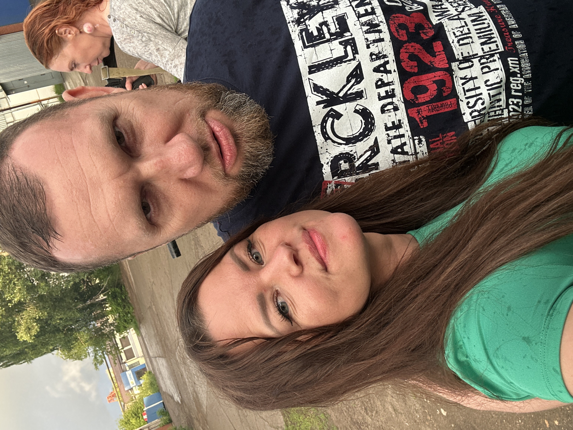 An outdoor, eye-level selfie is taken by a couple in front of a building with a gray metal facade in Chaikovsky, Russia. The man, in the foreground, has a beard and is wearing a dark blue t-shirt with white and red lettering. The woman, beside him, has long brown hair and is wearing a green t-shirt. In the background, a woman with red hair is visible, looking to the side and wearing a light gray shirt. The environment appears to be a street or an industrial area, with a building and some greenery visible.