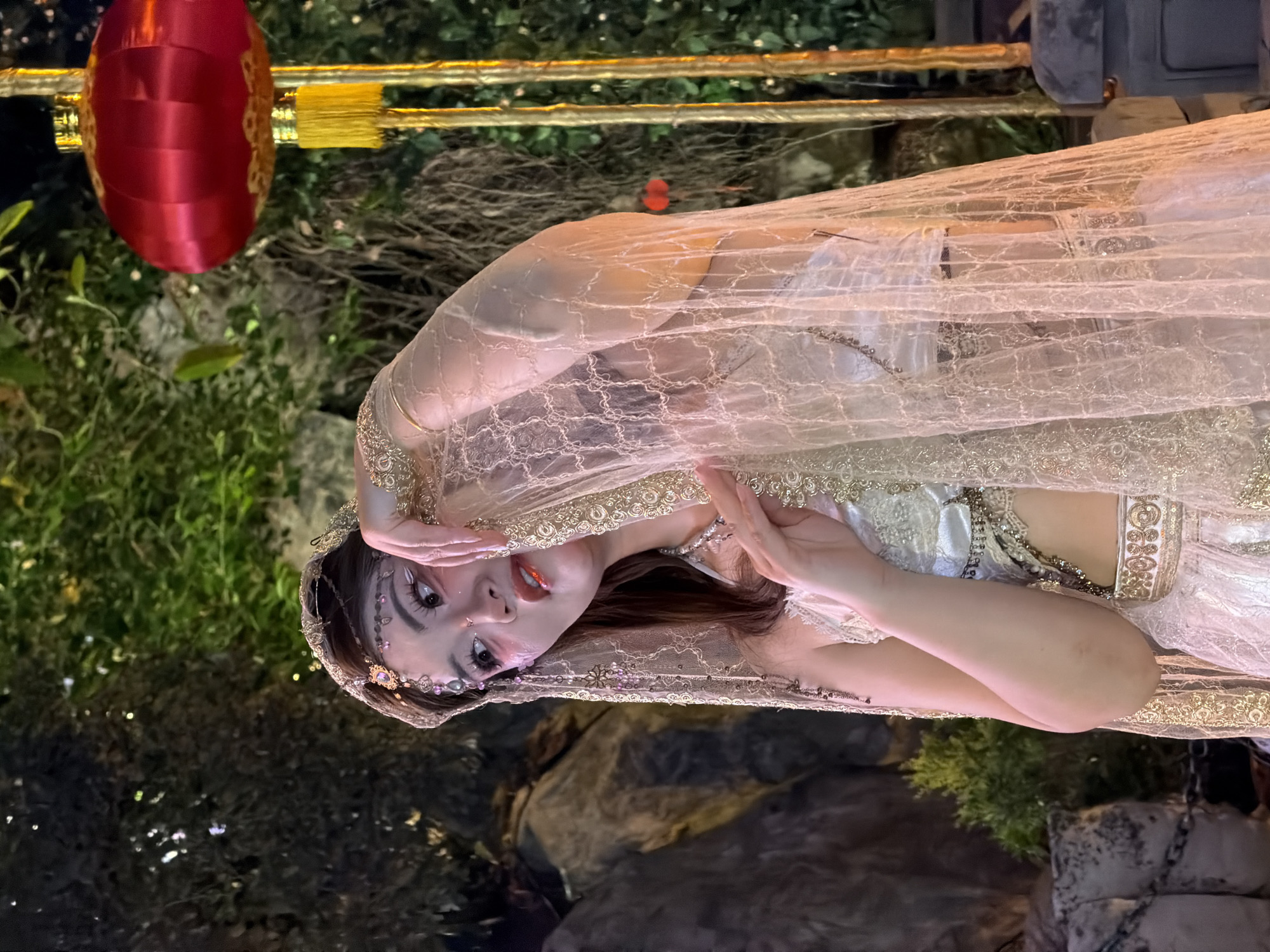 A person, dressed in ornate, shimmering attire with a veil covering their head and shoulders, is positioned in an outdoor setting. The person's face, adorned with dramatic makeup including defined eyes and bold lipstick, is visible as they look towards the camera. Their hands are gently placed near their face. The attire appears to be theatrical or ceremonial, featuring intricate embroidery and beading in gold and silver tones. The background suggests a garden or natural landscape with foliage and rocks, and hints of man-made structures like railings and possibly decorative lanterns, one red lantern being prominently visible in the upper left of the frame. The lighting suggests it might be evening or nighttime, given the presence of artificial illumination. The overall mood conveyed is one of fantasy or enchantment. There is no visible text in the image.