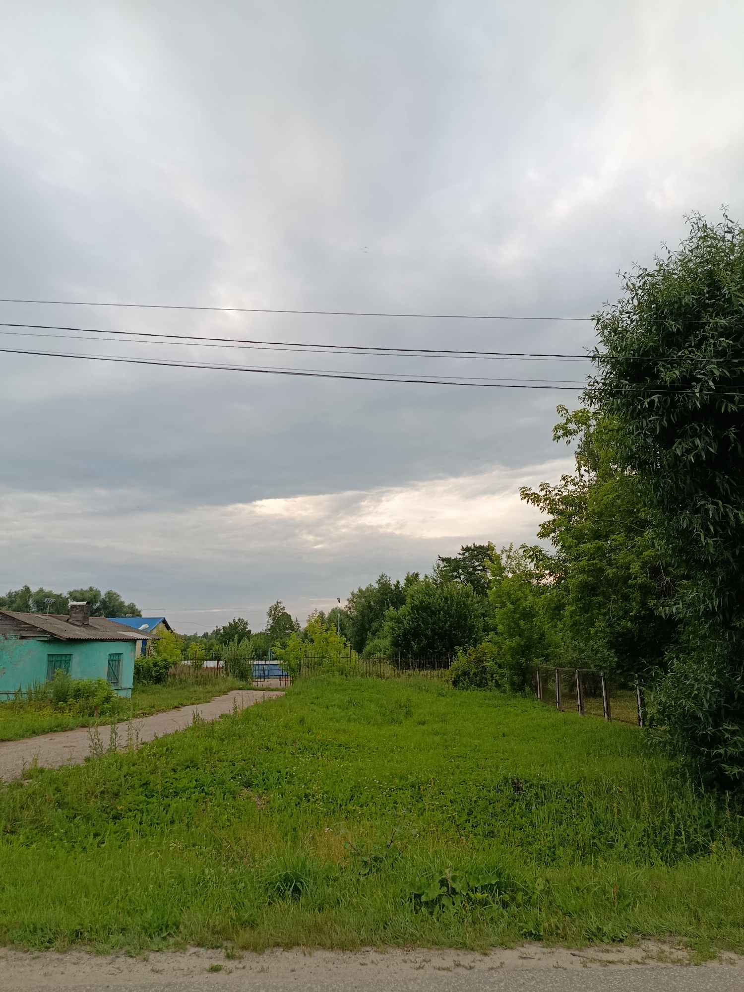 This media file depicts a rural scene in Verezya, Russia. The foreground is dominated by a grassy slope with a dirt path winding its way from the bottom left corner towards the center. Beyond the path, a small, weathered, teal-colored building with a dilapidated roof and boarded-up windows is visible on the left. Further in the background, trees line the horizon, with a few other buildings, including one with a blue roof, nestled among them. A metal fence with vertical posts runs along the right side of the frame, separating the grassy area from more dense vegetation. Overhead, three power lines stretch across the cloudy sky. The sky itself is overcast with patches of bright light suggesting the sun is attempting to break through. The overall impression is that of a quiet, somewhat rustic, and possibly overcast day in a rural Russian village. No people or specific events are depicted, and no text is visible. The time of day appears to be late afternoon or early evening, given the quality of light.