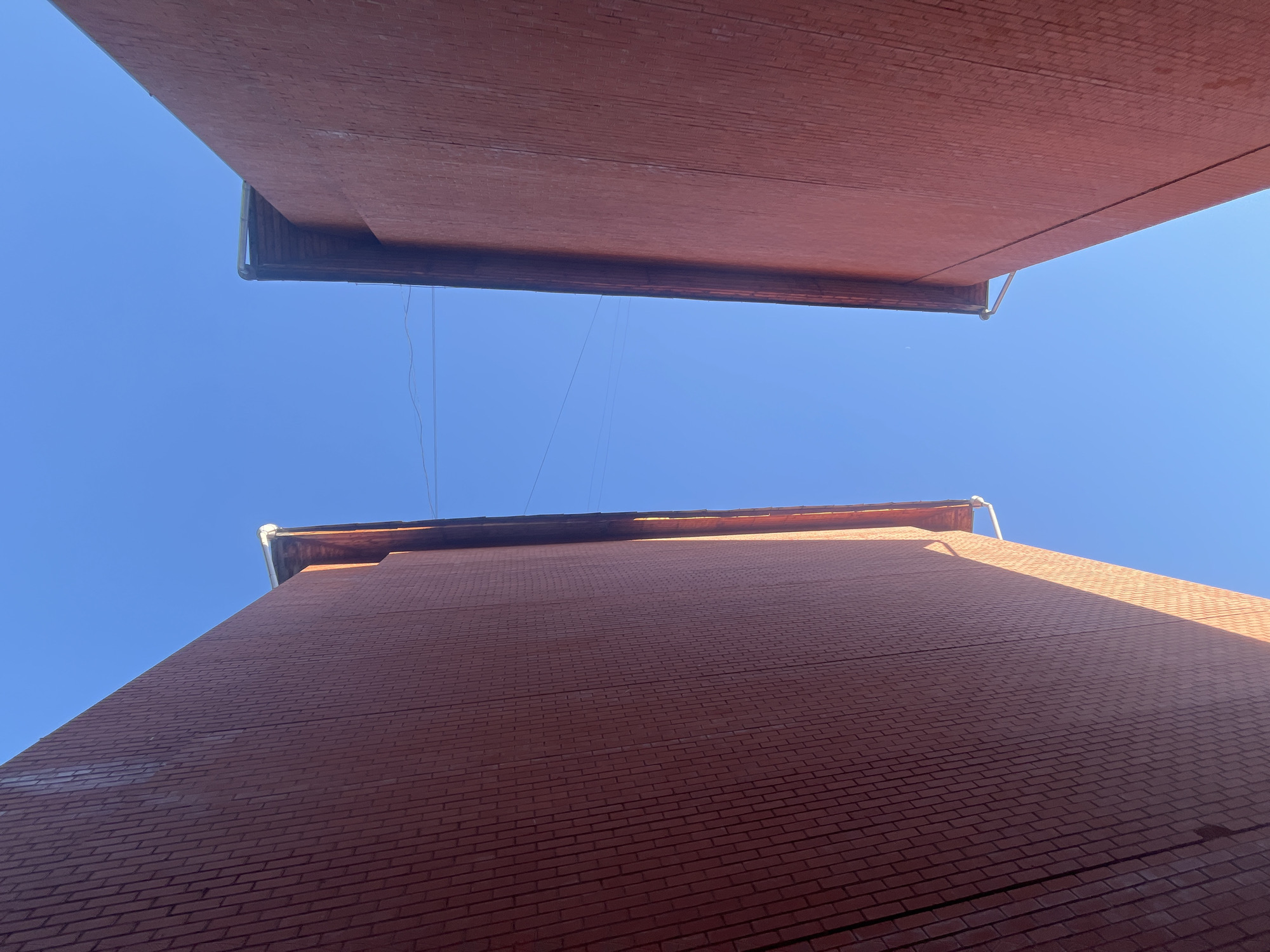The image shows a low-angle view looking up at two brick buildings against a clear blue sky. The buildings appear to be very close together, with the top of one building and the underside of another filling most of the frame. Both structures are made of reddish-brown bricks. One can see gutters attached to the roof edges of the buildings. Thin cables or wires are visible stretching between the buildings against the blue sky. The scene appears to be in an urban environment.