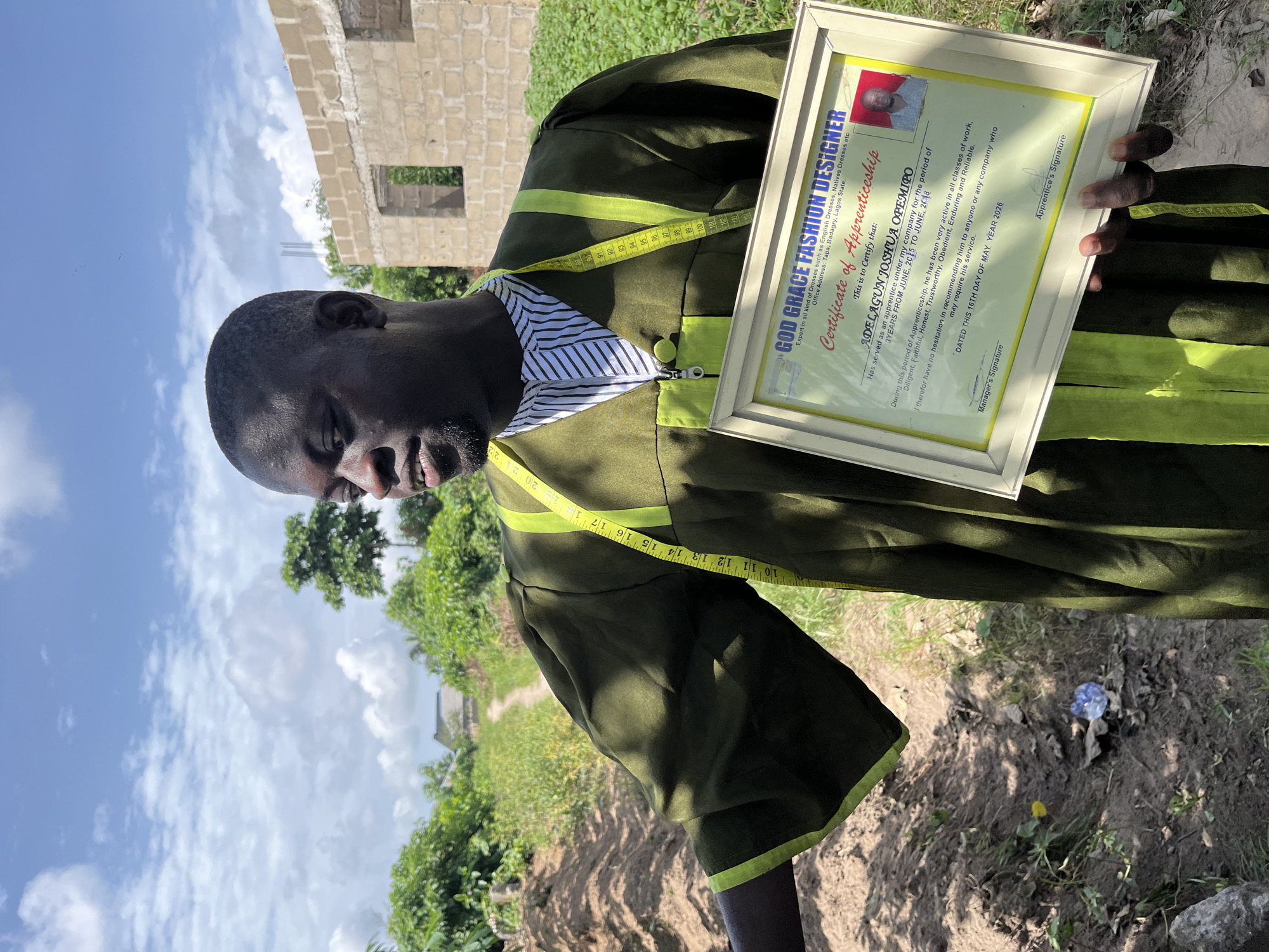 An African male stands outdoors, holding a framed "Certificate of Apprenticeship" from "God Grace Fashion Designer." He wears a green garment with neon yellow trim and a striped shirt visible at the collar. A measuring tape hangs around his neck. The certificate is dated May 16, 2026, and certifies that "ADELAGUN JOSHUA OPEMIPO" has served as an apprentice for three years from June 2015 to June 2018. A building under construction is visible in the background. The ground appears to be a mix of dirt and grass. The sky is partly cloudy. The certificate indicates the office address of the fashion designer is in Tapa, Badagry, Lagos State.