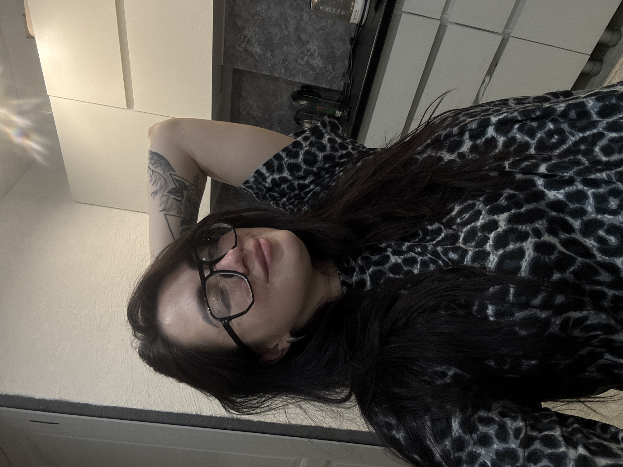 A medium close-up shot, rotated 90 degrees clockwise, features a person with long, dark hair and black-framed glasses lying down and looking towards the viewer. The person is wearing a black and grey leopard print top. A detailed tattoo, appearing to be a bird or mythical creature with floral and geometric elements, is visible on their upper left arm. Their expression is relaxed, with lips slightly parted.
The setting appears to be indoors, possibly a modern kitchen or living area. In the background, there are light-colored, possibly white, cabinet doors or wall panels. A dark, textured surface, possibly a countertop, is also visible with some indistinct items on it, including what looks like a small appliance or container. A bright light source or window causes a lens flare effect in the upper left corner of the rotated image. No discernible text is visible. The overall mood is casual and relaxed.