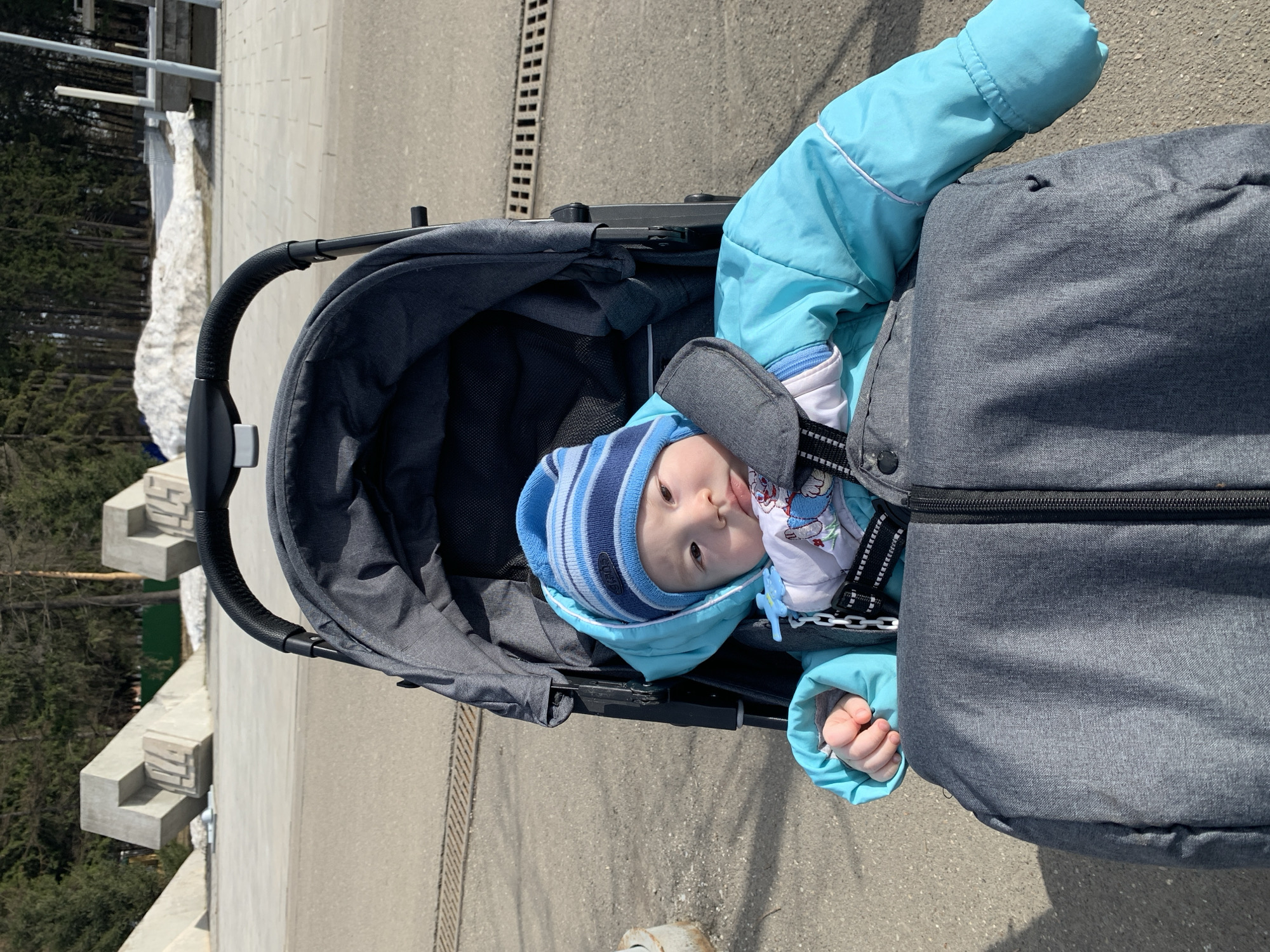 A young child wearing a blue and white striped hat and a blue winter coat is lying in a gray stroller. The stroller is on a concrete surface. In the background, there is a patch of snow near a concrete structure, possibly steps. Trees are visible in the distance. The location is likely Izhevsk, Russia.