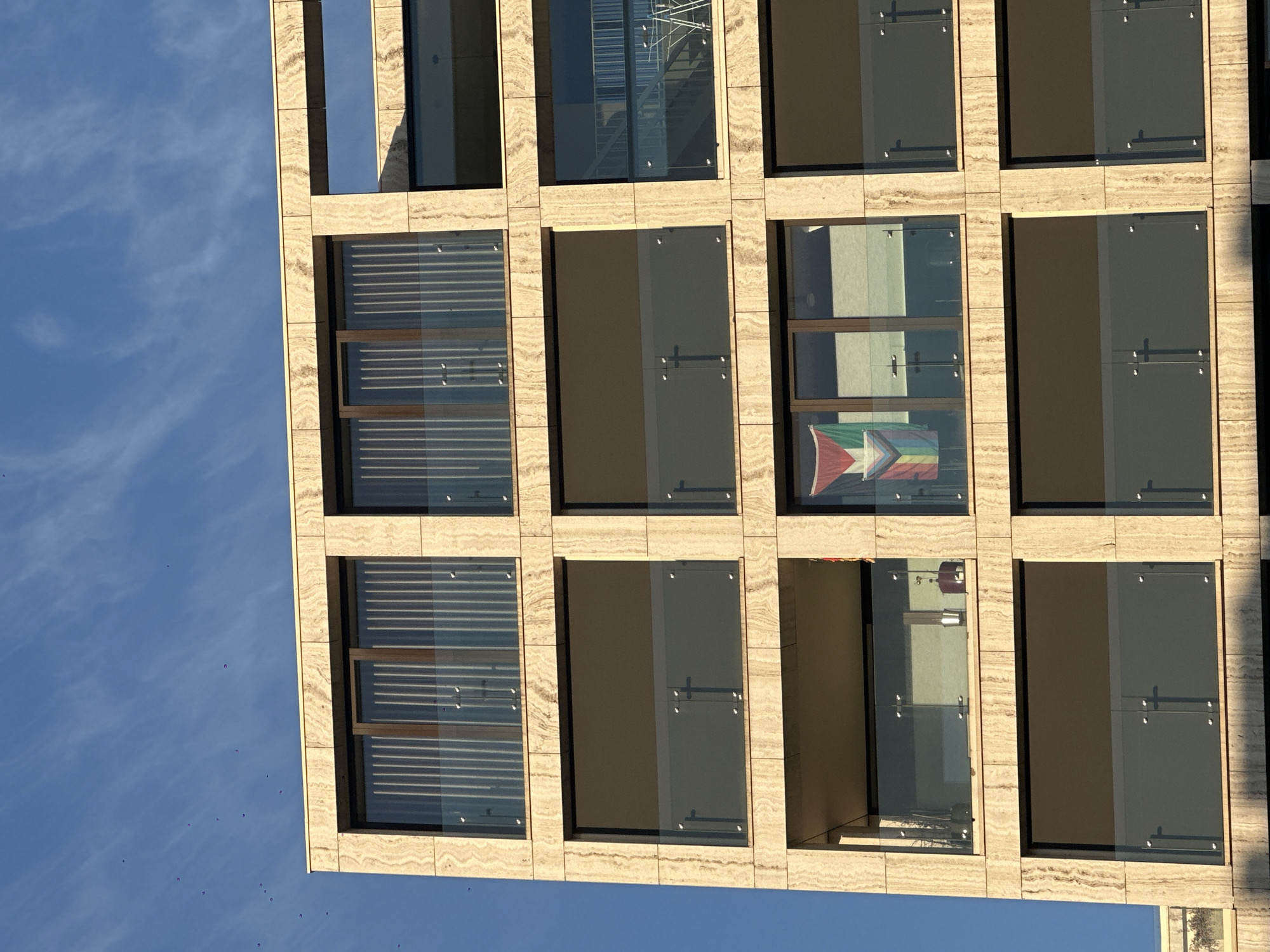 The image shows a modern apartment building with a light beige, textured facade. The building has multiple floors with large windows and balconies. On the third floor, a flag is visible hanging in one of the windows. The flag has horizontal stripes of black, white, and green, with a red triangle on the left side, and a smaller rainbow-colored chevron on the right. This flag appears to be a variation of the Palestinian flag with added LGBTQ+ symbolism. The sky above is blue with wispy white clouds. A few small birds are visible in the sky. The scene is likely captured in a city environment given the architectural style of the building.