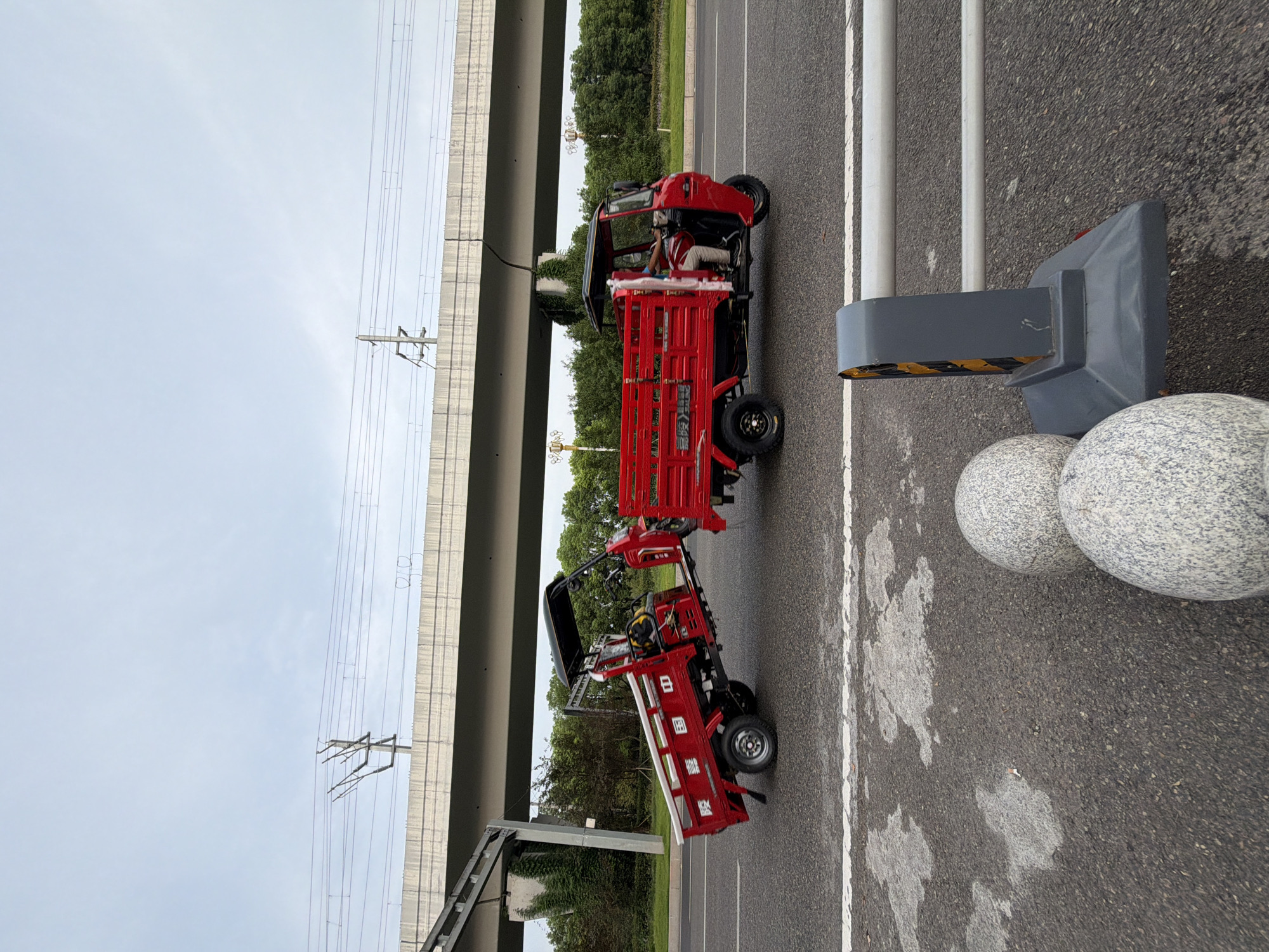 This is a medium shot, eye-level, outdoor, daytime photograph of two red, three-wheeled trucks on a paved road in Jiangjin, China. The trucks are parked side-by-side and are tethered together. The truck on the left is slightly in front of the truck on the right. Both trucks have open beds. The truck on the right has a driver seated in the cab. The background features a concrete overpass with electrical wires running across it, lush green trees, and a cloudy sky. In the foreground, there is a concrete barrier with white horizontal rails, and two large, speckled, spherical bollards.