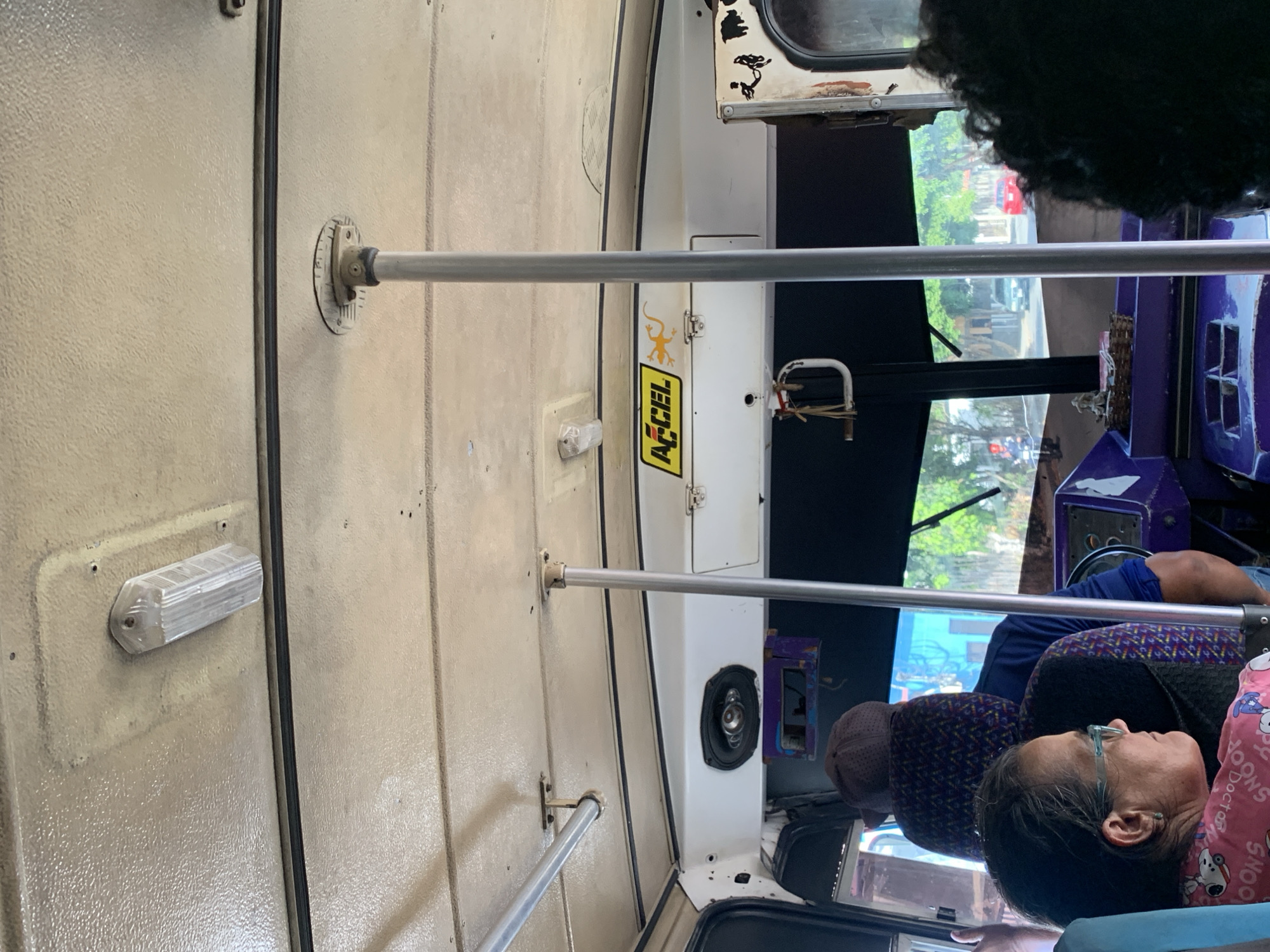 This image captures the interior of a public transport vehicle, likely a bus or large shared taxi, in La Asuncion, Venezuela, during daylight hours. The perspective is from a passenger's seat, looking towards the front and right side of the vehicle.
The interior features light beige, textured paneling on the ceiling and upper walls, with dark rubber or plastic trim lines separating sections. Silver-colored metal grab rails extend horizontally across the upper interior, supported by circular mounts, and another shorter rail is visible lower down. A clear, rectangular light fixture is attached to the beige paneling. Towards the front, a white panel separates the main passenger area, housing an embedded oval speaker and a small, white, D-shaped handle or latch with twine wrapped around it. On this white panel, a prominent yellow and black sticker displays the text "A-CEL," with a small yellow gecko graphic above it.
In the foreground, a person with dark hair and light-framed glasses is seen in profile, facing right. They are wearing a pink top patterned with "SNOOPY" characters and text. This individual appears to be a passenger, possibly resting. Behind them, part of another passenger's arm in a blue shirt is visible, and a dark, curly head of hair is partially seen in the upper right. A brown cap rests on a seat in front of the main subject.
Through the vehicle's windows, an outdoor urban or suburban scene is visible, showing green trees, buildings, a road, and other vehicles, including a red car. The bright light suggests a clear and sunny day. The driver's area is partially visible through the window frame, showing a purple-colored console or control unit.