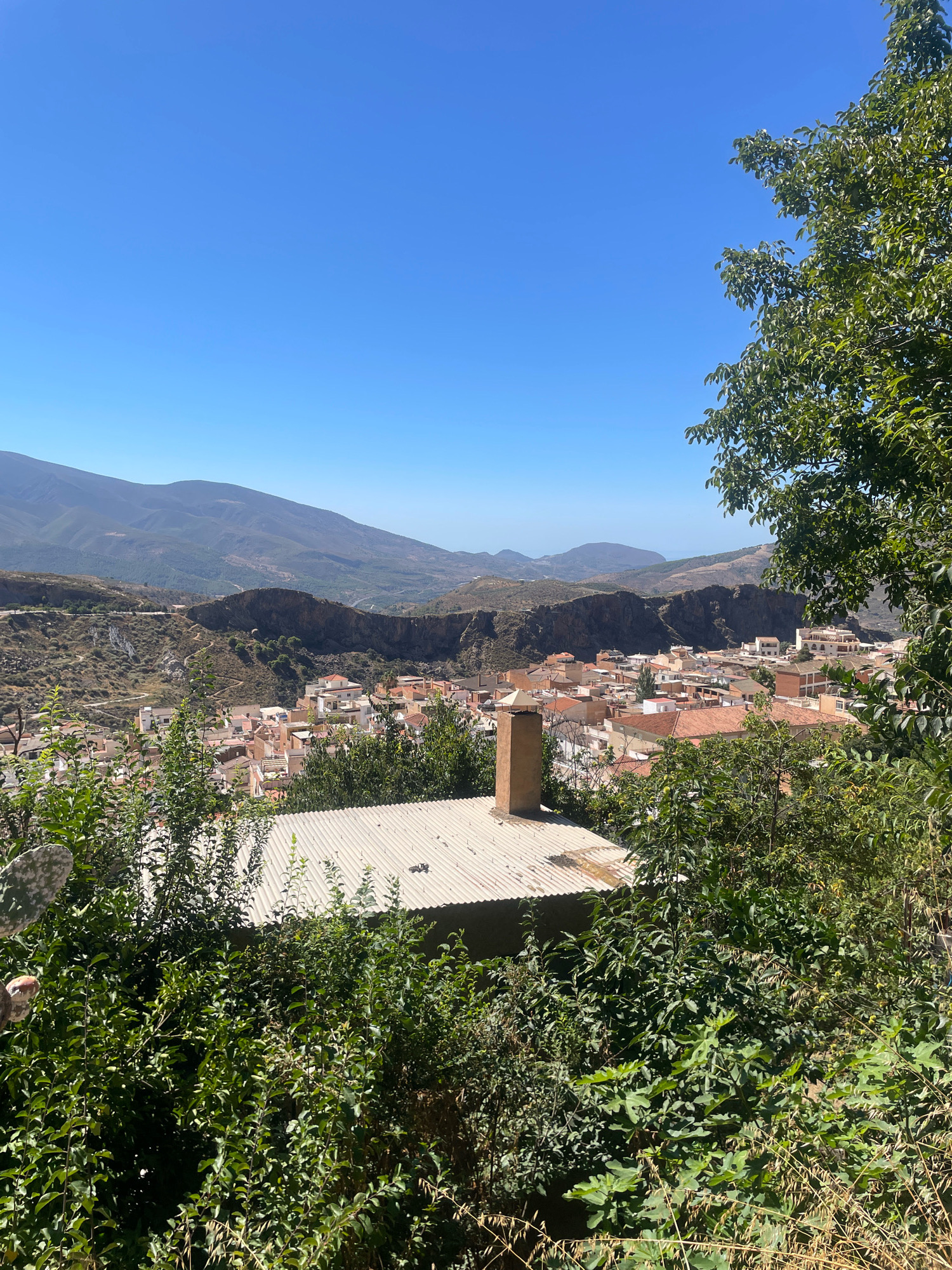 A high-angle, wide shot captures a sun-drenched Spanish village nestled in a valley. The sky is a clear, vibrant blue. In the foreground, lush green foliage and a portion of a corrugated metal roof with a chimney dominate the lower third of the image. Beyond this, a cluster of buildings with terracotta tile roofs forms the village. The village is set against a backdrop of arid, hilly terrain with winding roads. In the distance, large, hazy mountains stretch across the horizon. The scene is static and quiet, suggesting a peaceful afternoon in the Andalusian countryside.