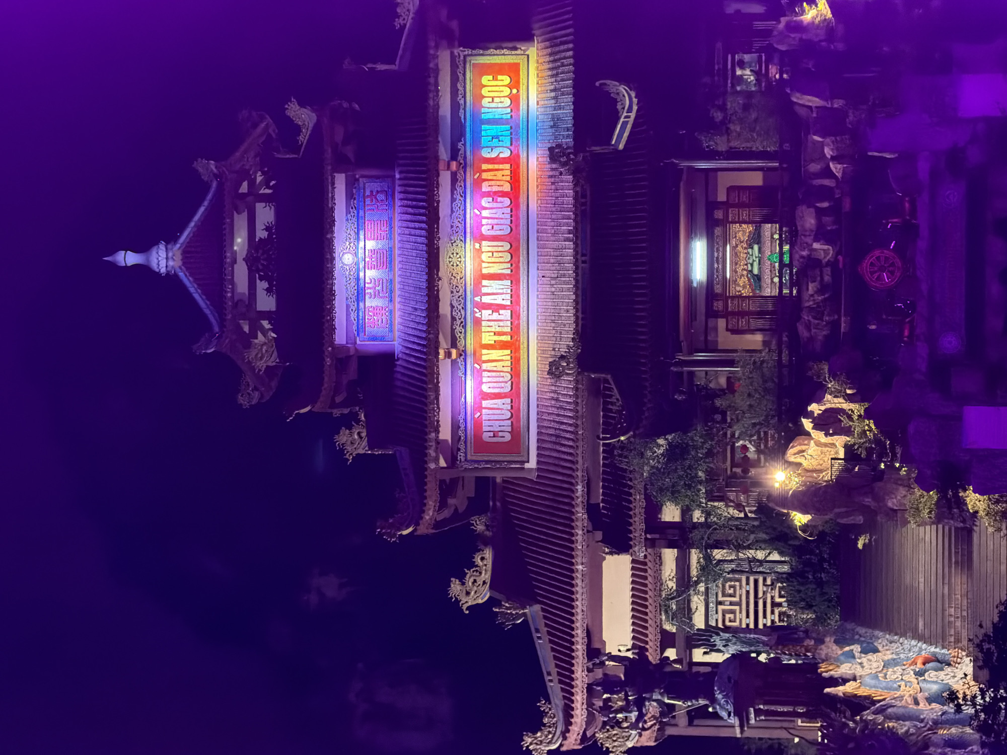 The image shows the Quan Thế Âm Buddhist temple in Đà Nẵng, Vietnam, at night. The architecture is traditional East Asian, with multiple tiers of tiled roofs and ornate carvings. Two vertically oriented signs are visible on the building, illuminated with bright, multi-colored neon lights. The sign furthest from the viewer displays Chinese characters. The closer sign has Vietnamese script, with the top portion reading "CHUA QUAN THE AM NGU GIAC DAI SEN NGOC". Additional details include decorative dragon sculptures and a well-lit courtyard with landscaping. The environment is dark, suggesting nighttime, with a purple tint likely from the lighting or image processing.