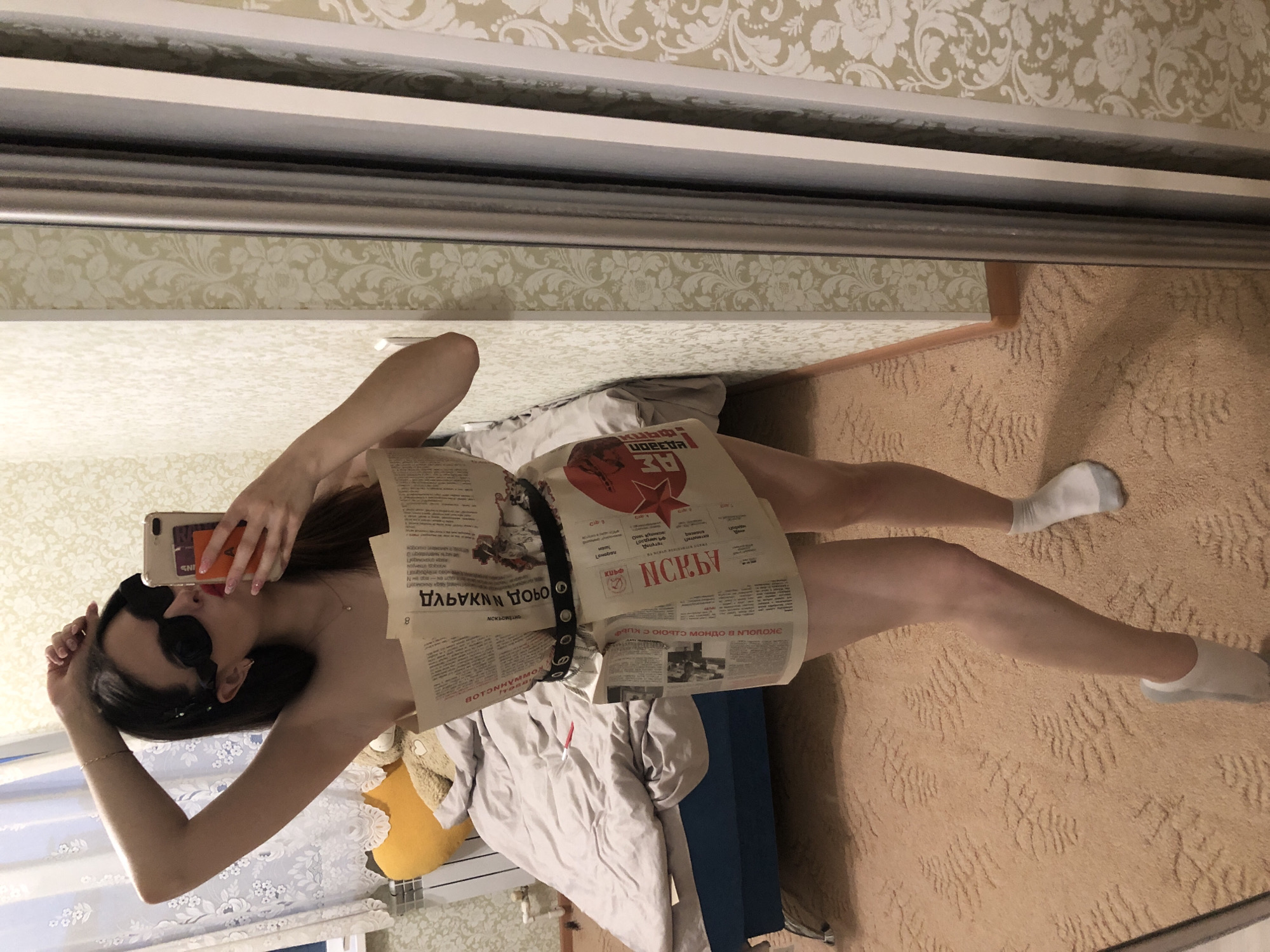 This mirror selfie captures a young woman standing indoors, dressed in an improvised outfit made of newspaper sheets, held at her waist by a black belt with silver eyelets. She wears dark sunglasses and white socks, holding a beige smartphone in her left hand to take the photo. Her fingernails are manicured pink.
The newspaper text is in Russian, with visible words such as "ИСКРЫ" (Sparks), "Победа" (Victory), and "ПОЛОЖЕ" (position). A prominent red star graphic is also seen on one of the newspaper sections.
The background reveals a room with light-colored, patterned wallpaper and a textured beige carpet. Behind the woman's legs, a light grey comforter or duvet is visible on what appears to be a bed, alongside a yellow, possibly animal-shaped pillow. A blue object, possibly furniture, is partially visible on the floor. The scene suggests an indoor setting, likely a bedroom or dressing area in Noviy, Russia, given the newspaper language and context.