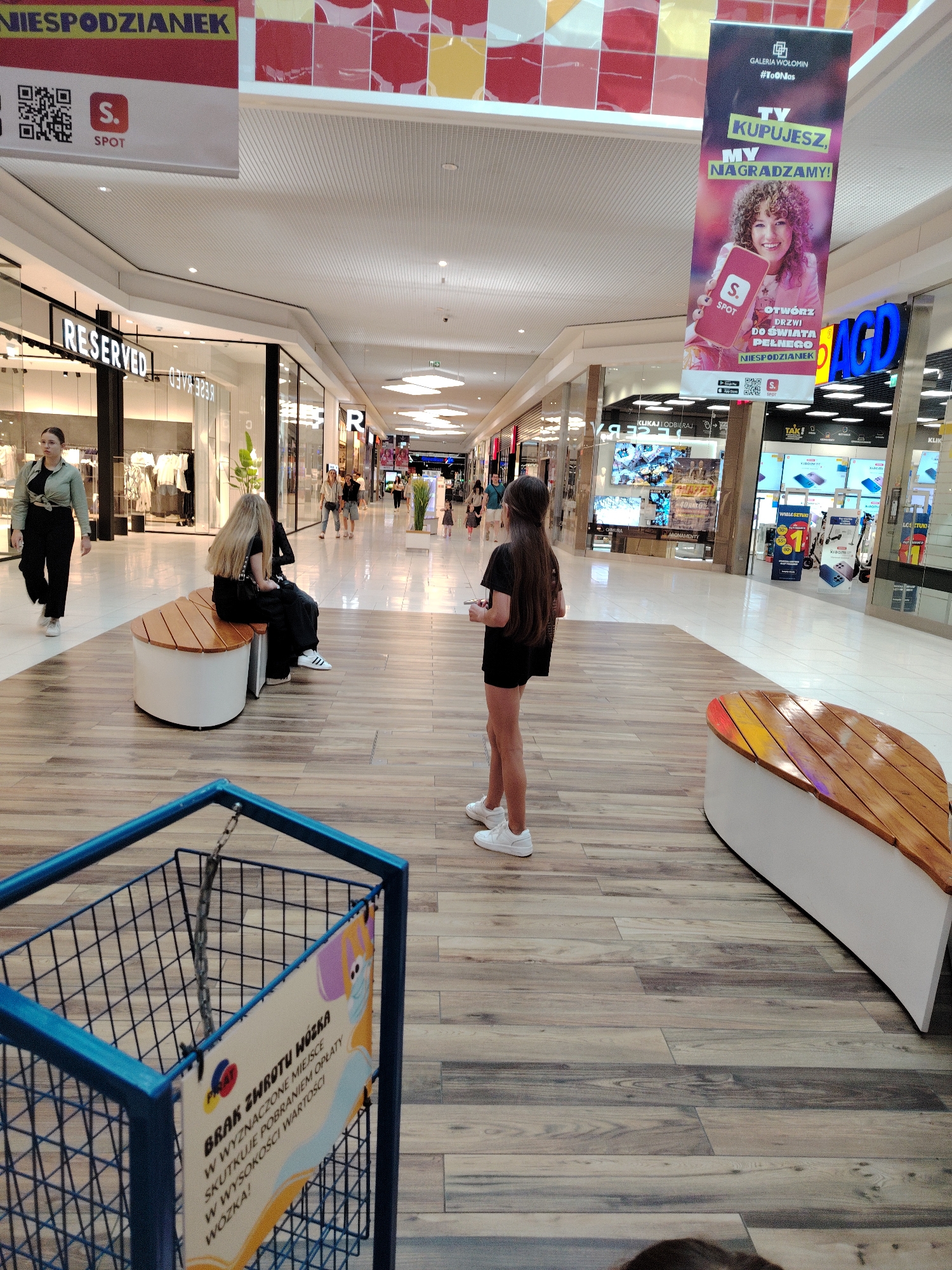 The image depicts the interior of a shopping mall in Wołomin, Poland. In the foreground, a blue shopping cart with a sign attached to it is visible. The sign displays text in Polish and an illustration of a cartoon character wearing a face mask. In the middle ground, two individuals are seated on a curved wooden bench. One has long blonde hair and is wearing dark clothing, while the other is obscured from view. Another individual, wearing a light green shirt and dark pants, walks away from the camera towards a store labeled "RESERVED." In the background, several shops are visible, including "RESERVED" and "AGD," which sells home appliances. Banners hang from the ceiling, one featuring an advertisement for "GALERIA WOŁOMIN" with the slogan "TY KUPUJESZ, MY NAGRADZAMY!" (You buy, we reward!). The mall has a modern design with a tiled floor and a suspended ceiling.