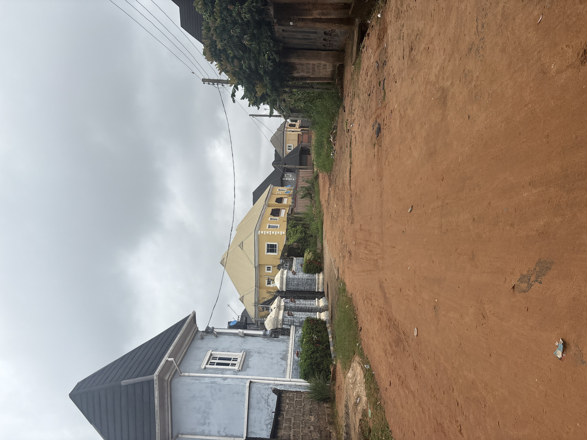 The image depicts a street scene in Obosi, Nigeria, characterized by its unpaved, reddish-brown dirt road. Several residential buildings line the road, showcasing a variety of architectural styles. On the left, a pale blue two-story house with a dark, steeply pitched roof and white trim stands prominently. Next to it, an ornate gate with grey and white pillars and black metal bars is visible, bordered by neatly trimmed green bushes. Further down the road, a large yellow house with a distinctive hip roof and white window frames is prominent. Other houses with similar roofing styles and muted color palettes are visible in the background, suggesting a developed residential area. Electrical wires and poles are strung across the scene, indicating the presence of utility infrastructure. The sky is overcast with grey clouds, suggesting a cloudy or potentially rainy day. The road itself shows tire tracks and scattered debris, including plastic wrappers, indicative of typical urban street conditions. There are no people or vehicles present in the frame.