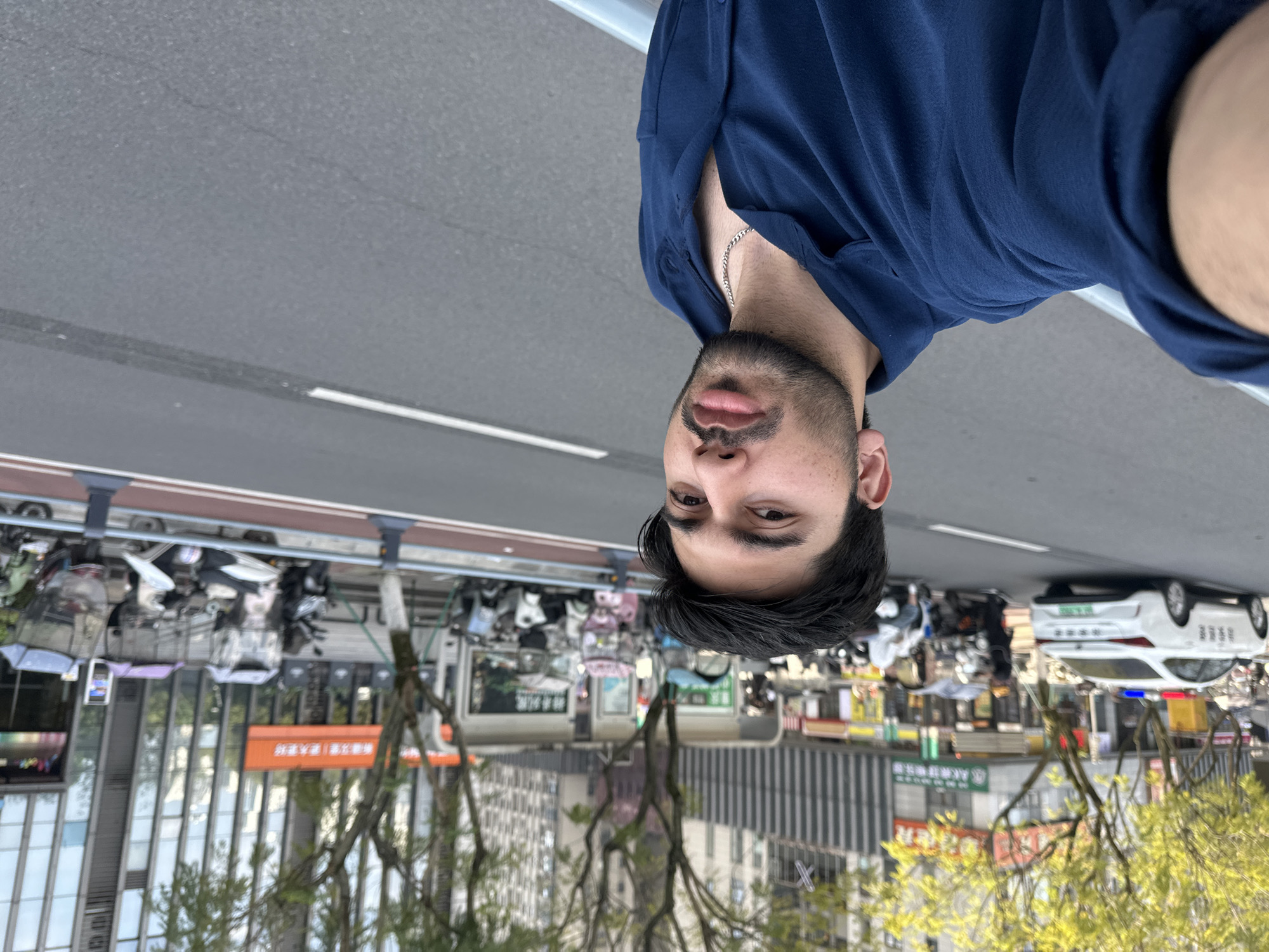 An overhead-style selfie captures a man from the chest up, oriented upside down. He has dark hair, a beard, and is wearing a dark blue collared shirt with a silver chain around his neck. His expression is neutral, looking directly at the camera.
The background, also oriented upside down, reveals an urban street scene from an elevated perspective, likely a bridge or overpass. A gray asphalt road with white lane markings is visible, with a line of parked scooters and motorcycles lining one side. Beyond the parked vehicles, various buildings with reflective windows and green trees are visible. A white car, possibly a police or service vehicle with blue and red lights on top, is parked further down the street.
The scene depicts a typical daytime cityscape in Chongqing High-tech Zone, China, as confirmed by visible text. An orange rectangular sign in the mid-ground clearly displays Chinese characters: "分界立交 | 重庆高新" (Fenjie Overpass | Chongqing High-tech). Other smaller signs on buildings also feature Chinese text, but are not legible due to distance and resolution. The overall lighting suggests it's daytime, possibly overcast, with soft shadows. No specific activity beyond general urban life is distinctly observable in the background.