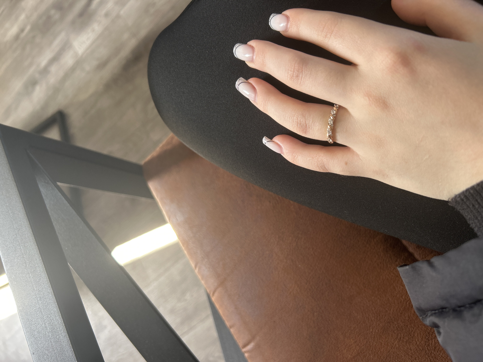 A close-up shot focuses on a person's left hand resting on a dark, possibly black, fabric surface. The hand features a ring on the ring finger, appearing to be a band of small, possibly diamond-like, stones set in a metal, possibly gold, setting. The fingernails have a light base with a fine black line following the curve of the nail tip, creating a minimalist, stylized French manicure effect. The hand is positioned over a brown leather-like cushion, with a section of a black metal chair frame visible in the periphery of the image. The backdrop is a light grey, possibly a wall or floor. The image does not provide information about location, including Kemul, Russia.