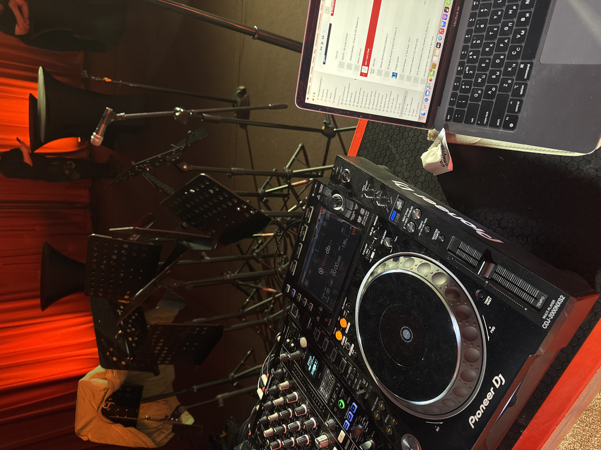 An overhead view captures a DJ setup and stage area, likely at an indoor event in Khimki, Russia. The scene is dominated by professional audio equipment, including a Pioneer DJ CDJ-2000NXS2 multi-player and mixer, connected to a MacBook Air laptop. The laptop screen displays music library software with various songs listed, some in English (e.g., "MALFA - So Long", "Gnarls Barkley - Crazy", "Calvin Harris - How Deep Is This Love") and some in Russian. The laptop keyboard shows Cyrillic characters, confirming the Russian context. A crumpled piece of white paper or napkin with the word "Спиртова" (Spirtova, likely "alcoholic" or related to spirits) is visible next to the laptop.
In the background, a series of dark music stands are arranged, suggesting live musicians or speakers may also be part of the event. A microphone is visible attached to a stand. Behind the stands, large, horn-shaped speakers are positioned. The backdrop consists of rich red curtains or drapery, illuminated to create a warm, atmospheric glow. A person's lower body, wearing light-colored attire, is partially visible among the music stands, and another person is partially visible in the far upper left. The overall setting indicates preparation for or the midst of a performance or gathering involving music. The DJ equipment has various labels like "rekordbox," "ECHO," "TEMPO," "BEAT SYNC," and "VINYL ADJUST." The laptop screen shows menu items such as "Музыка" (Music), "Артисты" (Artists), and "Плейлисты" (Playlists), with song durations and playback controls.