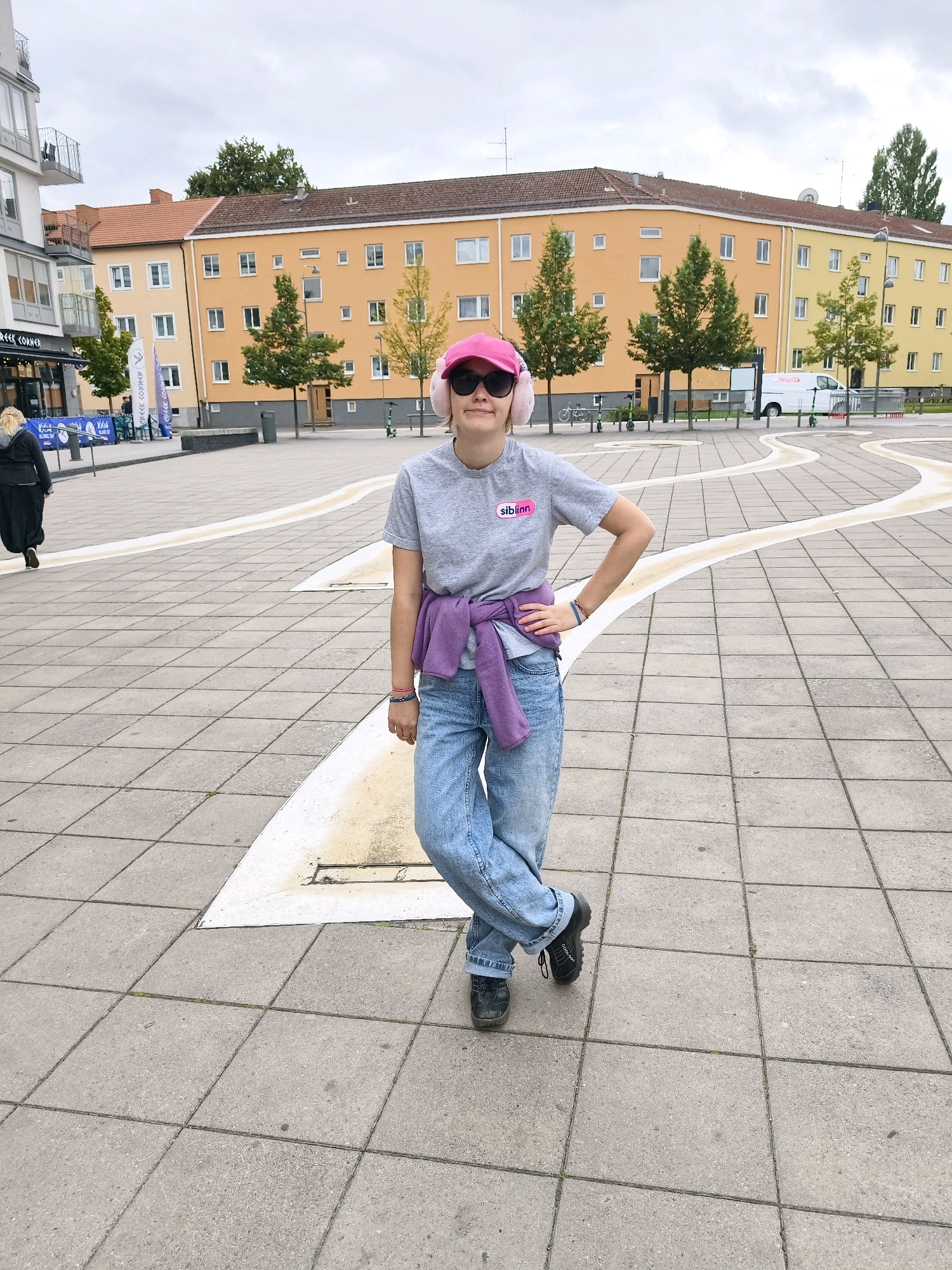 The image shows a young woman posing in a city square in Linköping, Sweden. She is wearing a grey t-shirt with a pink logo, light blue baggy jeans, and black shoes. A purple sweater is tied around her waist. She has on a pink baseball cap and pink earmuffs, and sunglasses. Her left hand is on her hip, and her right leg is crossed over her left.
Behind her, there is a street with buildings, trees, and a white van. The ground is paved with square tiles. In the background, people can be seen walking and standing. The overall setting is an urban environment with a mix of modern and older architecture. The sky is overcast.