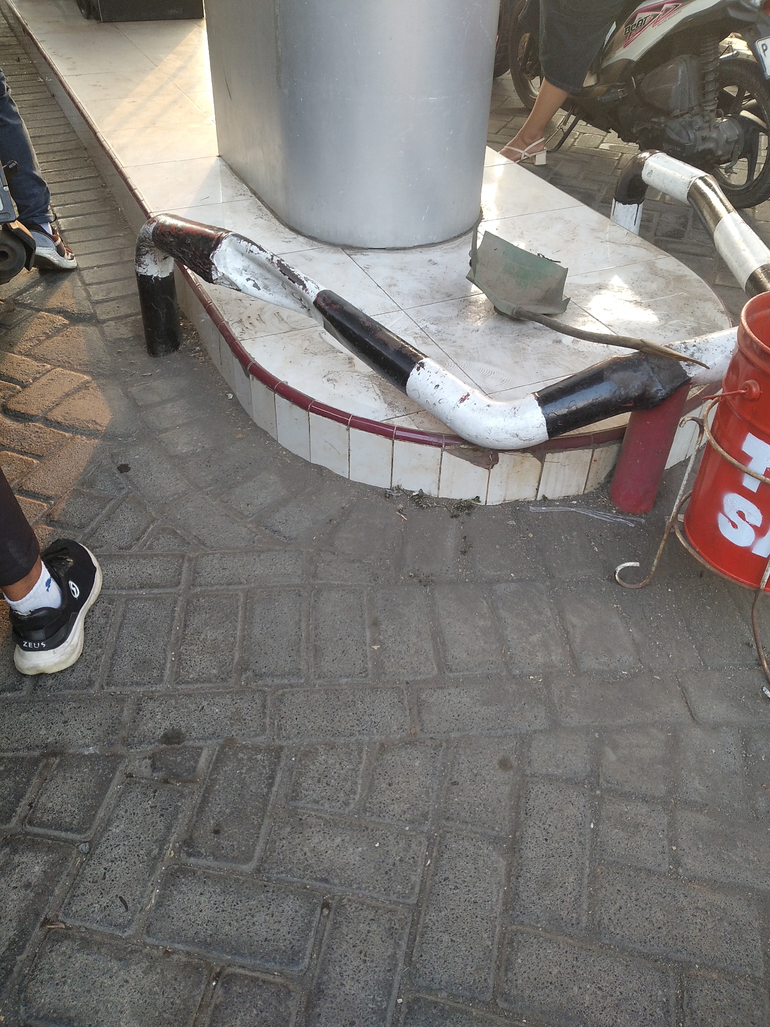 In Jubung, Indonesia, a section of a paved walkway features a large, gray, cylindrical pillar. A metal pipe, painted with alternating black and white stripes, is bent and secured to the ground near the pillar. The pipe appears to be part of a protective barrier or railing. To the right of the pipe, a red bucket sits on a metal stand. In the background, a portion of a motorcycle is visible, with a person's legs also in view, suggesting the area may be a parking space or thoroughfare. The walkway is constructed of dark gray interlocking rectangular pavers. In the foreground, a person's left foot, clad in a black sneaker with white laces and a white sole, is positioned on the pavers. The sneaker has "ZEUS" written on the heel. The lighting suggests it is daytime, with sunlight casting shadows on the walkway.