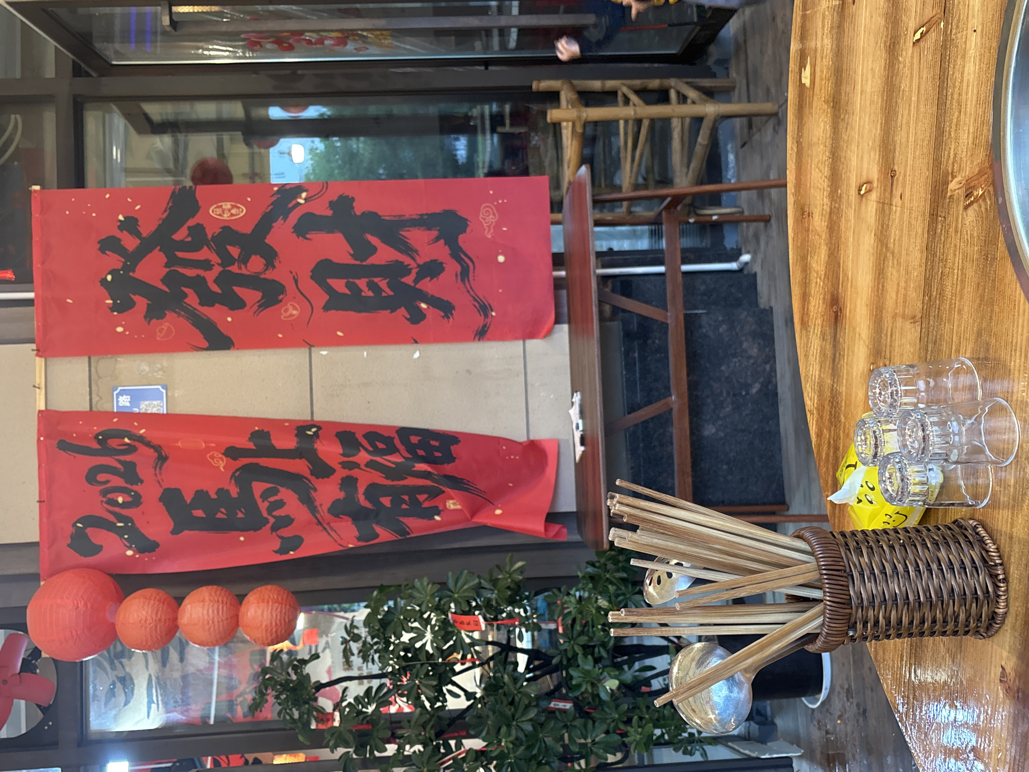 The image depicts an outdoor scene in Jiangjin, China, showing the exterior of a restaurant. Two red banners with black Chinese calligraphy hang above, possibly advertising the establishment or conveying a greeting. Below the banners, four red lanterns are visible. A small green plant is positioned in front of the window. To the right of the frame, a wooden table displays glasses, a yellow tissue pack, and a wicker basket containing wooden utensils, including ladles. In the background, part of a wooden structure resembling scaffolding is visible. A person's hand is briefly visible at the top of the frame.