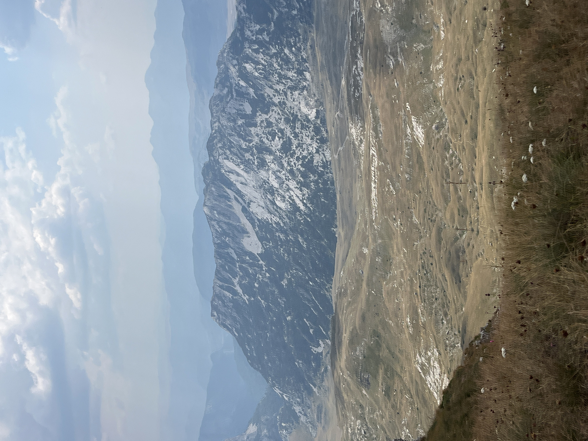 The image depicts a vast, mountainous landscape under a hazy, cloudy sky. The foreground is dominated by dry, tall grasses and scattered white and dark wildflowers, suggesting an arid or dry season. A steep, grassy incline leads the eye down into a wide valley. In the midground, the terrain transitions to rolling hills covered in dry, light brown and yellowish vegetation, with patches of lighter soil or rocks breaking up the surface. A winding dirt path is faintly visible in this area. The background is characterized by a large, imposing mountain range with rugged, rocky peaks. The mountains exhibit a mottled appearance of dark rock and lighter patches, possibly snow or lichen. Further in the distance, multiple layers of mountains fade into a pale blue haze, indicating significant depth and atmospheric perspective. The overall scene is natural and undeveloped, likely a remote wilderness area. Given the context of Боричје, Montenegro, this image likely represents a view within the Durmitor National Park, known for its dramatic mountain scenery and glacial valleys. There are no people or man-made structures clearly visible in the scene, apart from the faint path in the midground.