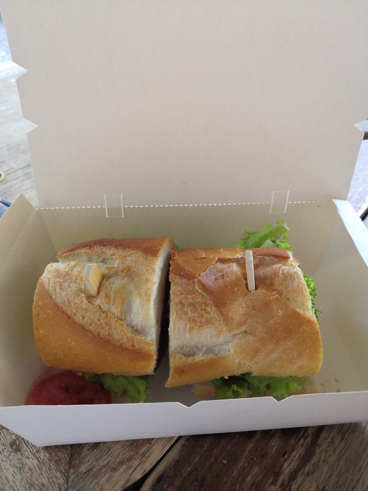 A close-up shot shows a baguette sandwich cut in half, held together with toothpicks, sitting in an open white cardboard takeout box. The sandwich contains lettuce and what appears to be tomato. The box is sitting on a wooden table. The location is Nha Trang Ward, Vietnam.