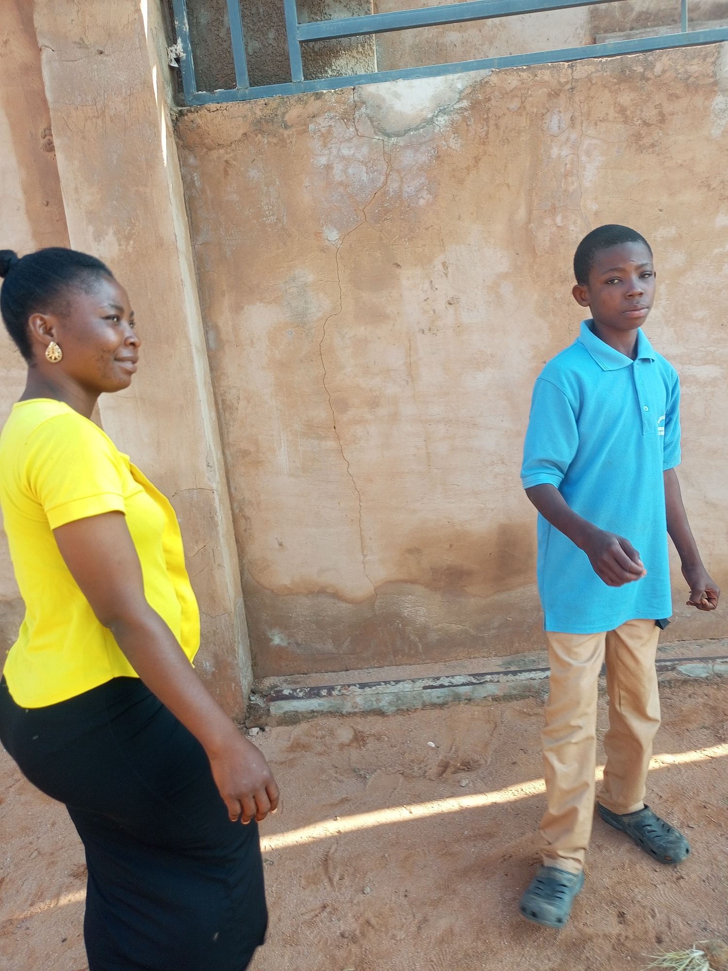 In Tempure, Nigeria, a woman in a bright yellow t-shirt and black skirt stands in profile, looking to the left. She has her hair pulled up in a bun and is wearing large, ornate gold earrings. Beside her, a young boy in a blue polo shirt and tan pants stands facing the camera, looking slightly to his right. He is wearing dark grey Croc-like shoes. They are standing on sandy ground in front of a weathered, textured wall with a dark metal grate structure above it. The lighting suggests it is daytime, with strong shadows cast on the ground.