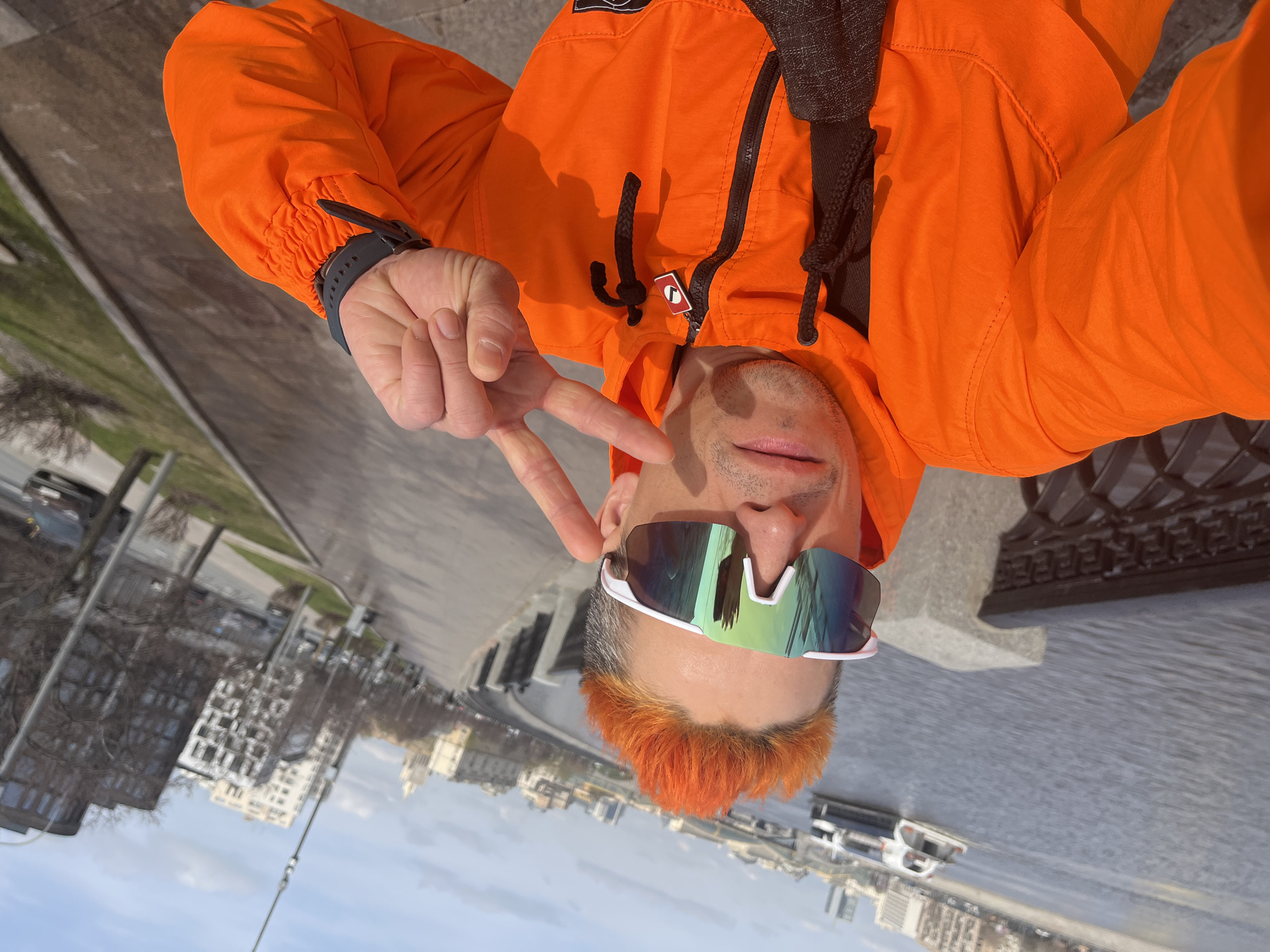 The image depicts a person in an orange jacket making a peace sign and wearing sunglasses. They have orange hair. The background reveals a city landscape, including buildings and a river. The location is the Moskva River embankment in Moscow, Russia.