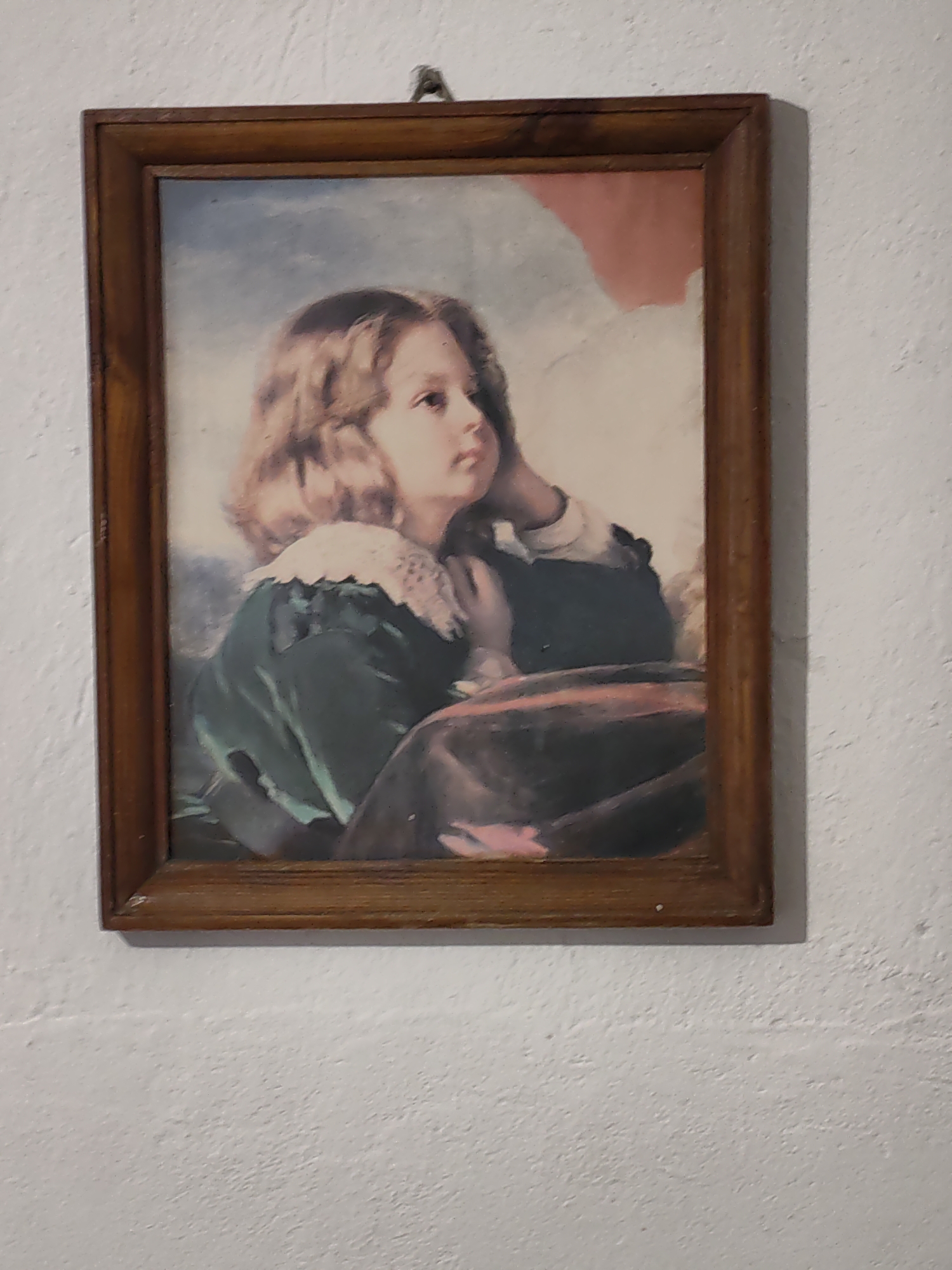 A framed portrait hangs on a textured, off-white wall. The frame is brown wood. The portrait appears to be a painting of a young child with light brown, wavy hair and fair skin. The child is looking off to the right of the frame. They are wearing a green garment with a white, lacy collar. Their left hand is visible against their cheek, and they appear to be resting on a dark brown or black object. A hint of pink or red color is visible in the background of the portrait. The location is Damianovo, Bulgaria.