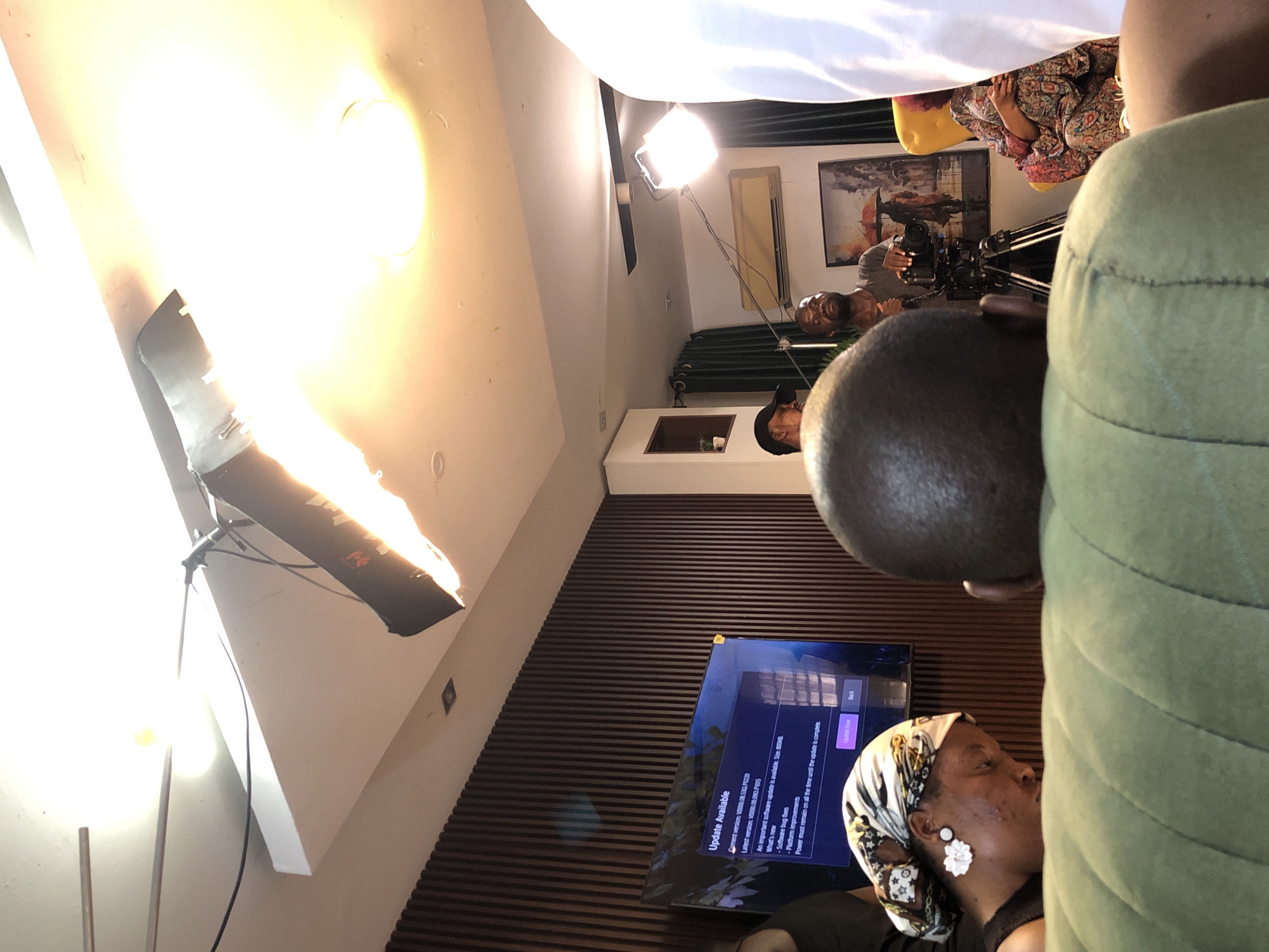 A room's interior features a television screen displaying an "Update Available" notification. Several people are present, including one wearing a headscarf with a white flower detail. A camera and lighting equipment are set up, suggesting a video or photo shoot. The location is Unknown, Unknown.