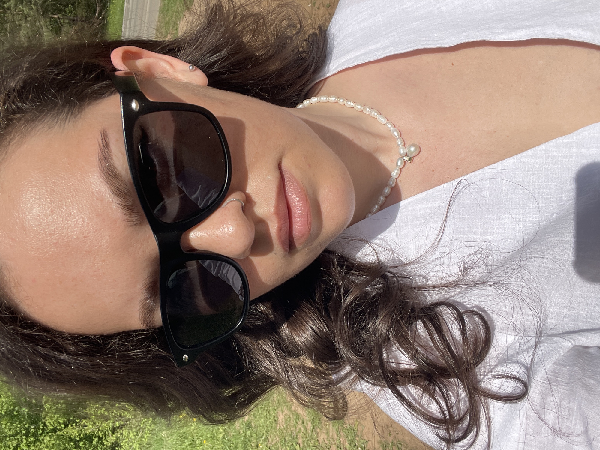 A woman lies on her side, outdoors, in a grassy area. She wears a white shirt, a pearl necklace, and black sunglasses. She has brown hair and a nose ring. The exact location in Izhevsk, Russia, cannot be determined from the visual information provided.