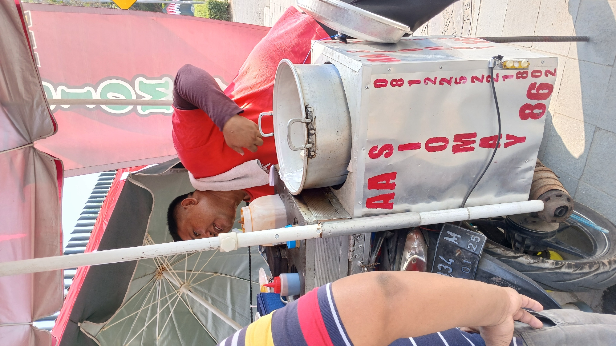 A man, wearing a red t-shirt and a towel around his neck, stands behind a food cart. The cart is primarily made of metal and is mounted on a motorcycle. Red lettering on the cart reads "AA SIOMAY" and a phone number "0812225221802". A large metal pot sits on top of the cart. The man appears to be interacting with a customer, whose arm and torso, wearing a striped shirt, are visible on the left side of the frame. The background suggests an outdoor urban environment, possibly a street or sidewalk in Borobudur, Indonesia, with a building and vegetation visible in the distance. The scene is captured in daylight.