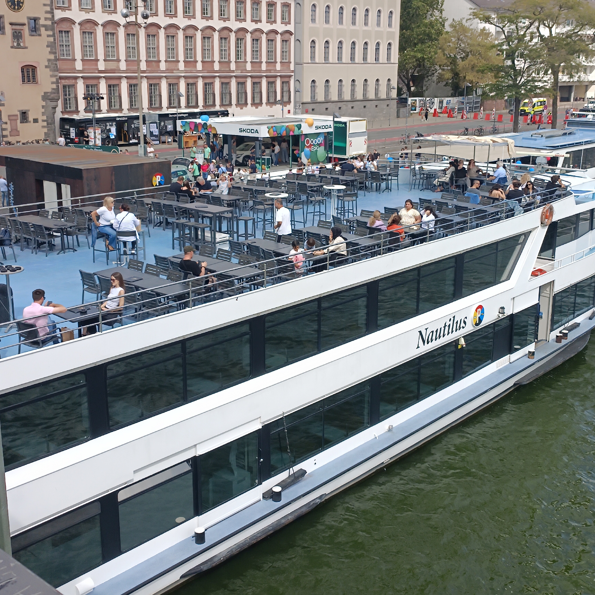 The image shows a modern white river cruise ship named "Nautilus" docked on the Main River in Frankfurt, Germany. The ship has multiple decks with large, dark-tinted windows. The upper deck is visible, featuring outdoor seating areas with numerous tables and chairs, occupied by many people. Some individuals are sitting, others are standing and conversing. The ship's name and logo are prominently displayed on its side.
Behind the ship, a street scene is visible with buildings of varying architectural styles. Some buildings appear older with ornate facades, while others are more modern. There are also several stalls and temporary structures set up along the waterfront, some with signs for "SKODA." People are seen walking around these stalls and near the water's edge. The overall atmosphere suggests a public event or a busy tourist area. The water of the Main River is a murky green color, and the weather appears to be clear.