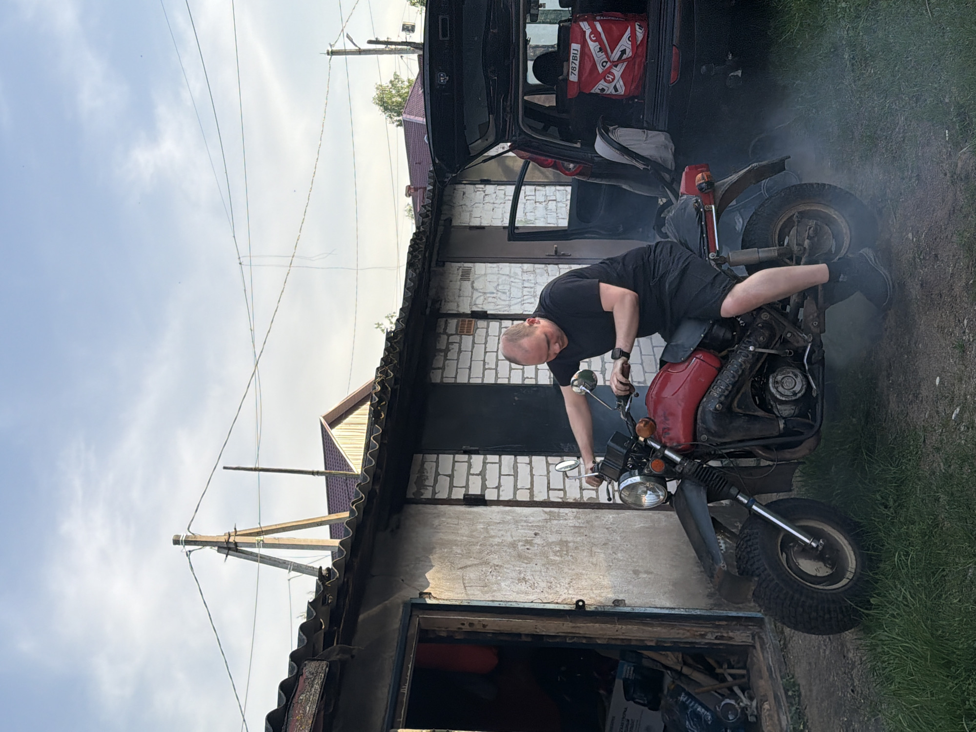 A man in black clothing sits on a red motorcycle, seemingly attempting to start it. Smoke is billowing from the rear of the motorcycle. The scene appears to be set outside a building, possibly a garage or shed, with a brick facade and a corrugated metal roof. A black SUV is partially visible in the background. The surroundings suggest a somewhat rural or semi-urban environment, and the presence of utility poles and overhead wires indicates some infrastructure. There is vegetation visible on the right side of the image, further suggesting a location outside of a densely populated urban area. The location provided, Reshetnikovo, Russia, supports this interpretation.