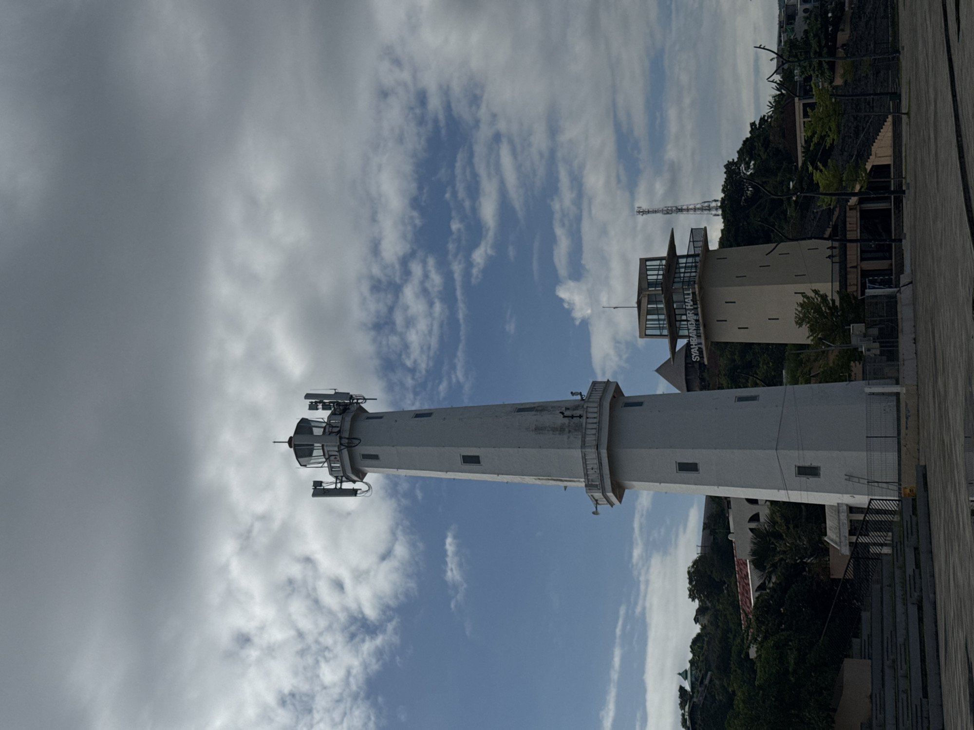 The image shows a tall, white lighthouse under a partly cloudy sky. The lighthouse has multiple levels and antennas at its top. Adjacent to the lighthouse is a building with a glass-enclosed observation area and the words "SYAHBANDAR HALL" on its side. The buildings are located on a hill with lush greenery. The foreground shows a paved area. This scene is located in Labuan Bajo, Indonesia.