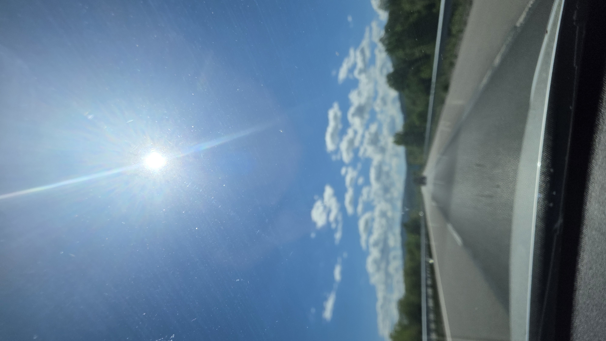The image shows a view from inside a vehicle traveling on a highway. The scene includes a clear blue sky with the sun shining brightly and some scattered white clouds. Alongside the highway, there are trees and greenery. The location appears to be a road in Blajievo, Bulgaria.