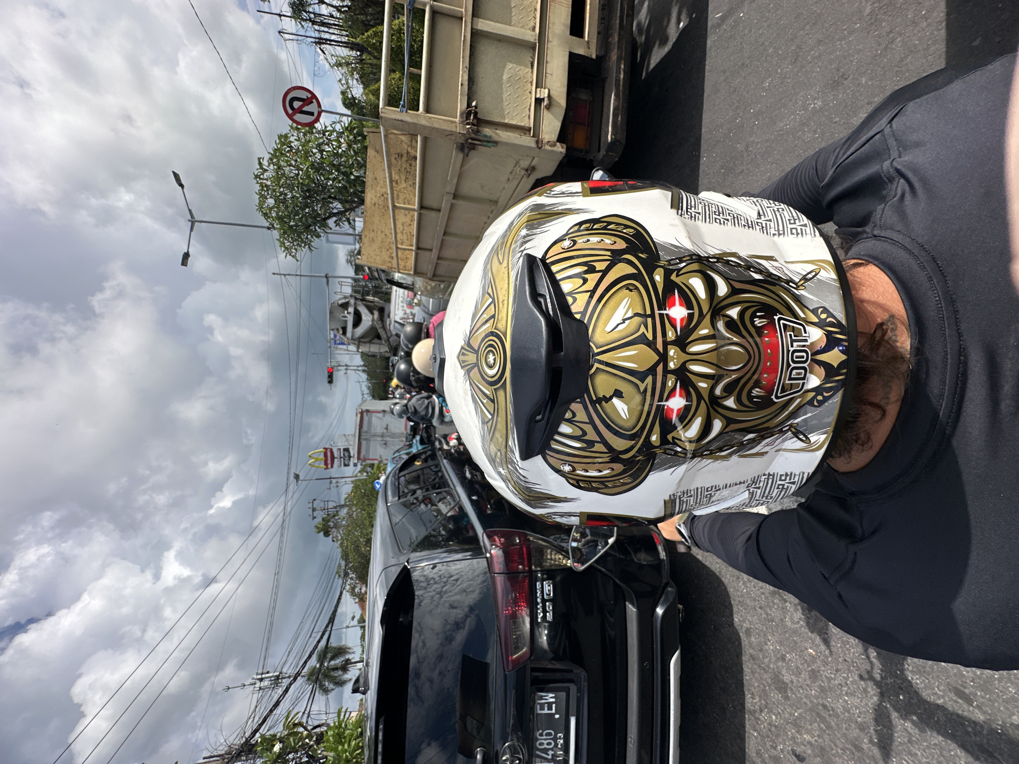 The image shows a motorcyclist's view while in traffic in Kuta Selatan, Indonesia. The motorcyclist wears a white and gold patterned helmet with "DOT" printed on it. They are stopped behind a black Toyota car with license plate "DK 1486 EW". Beyond the car, traffic is congested with other motorcycles and a truck. Street signs are visible, including a "No U-Turn" sign and a McDonald's restaurant sign. The sky is overcast with cloudy weather.