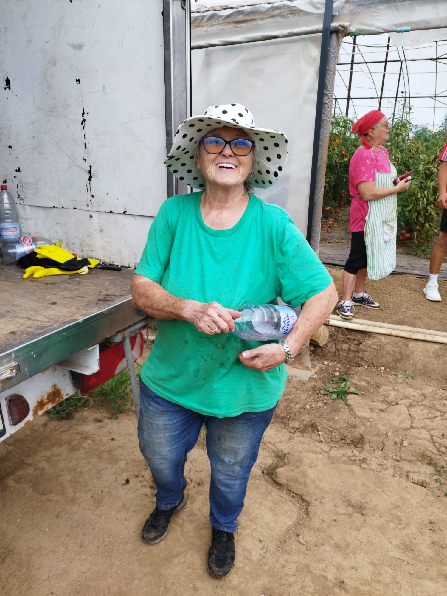In Băleni-Români, Romania, a woman wearing a green t-shirt, blue jeans, black shoes, and a white wide-brimmed hat with black polka dots stands outdoors. She is smiling and holding a plastic water bottle in her right hand. Her left hand is visible, and she wears a wristwatch and a ring. The background includes a white truck, a greenhouse with tomato plants, and another woman in a pink shirt and green and white striped apron, holding a red object. The ground is dirt.