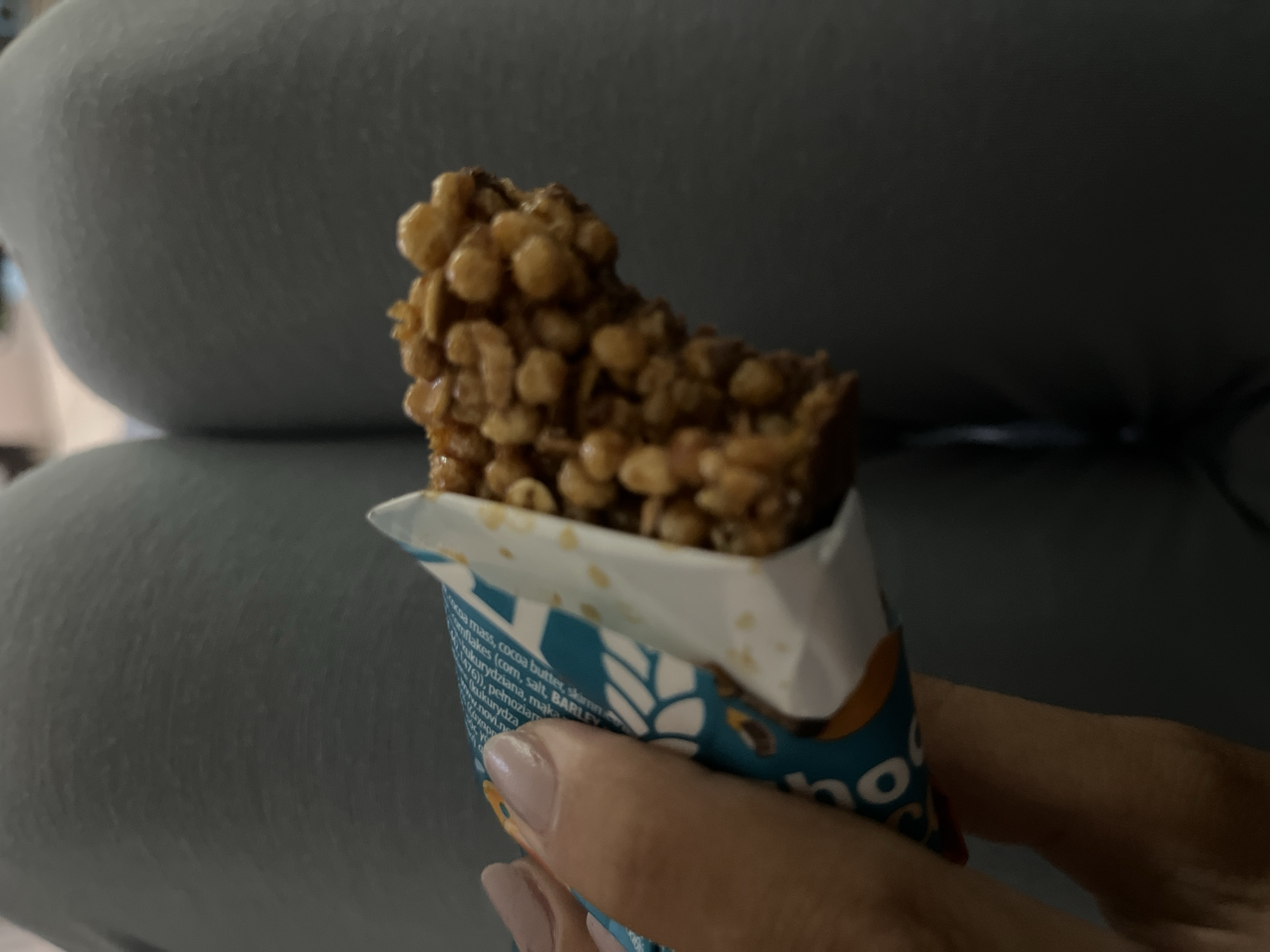 A person's hand is holding a partially unwrapped granola bar. The granola bar is rectangular, with a visible portion displaying a puffed grain texture, likely bound together by a sticky substance. The wrapper is blue and white with text in an unidentified language. Part of the ingredients list is visible, including "cocoa mass", "cocoa butter", "BARLEY", and "kukurydziana". The background is blurry but appears to be a blue upholstered surface.