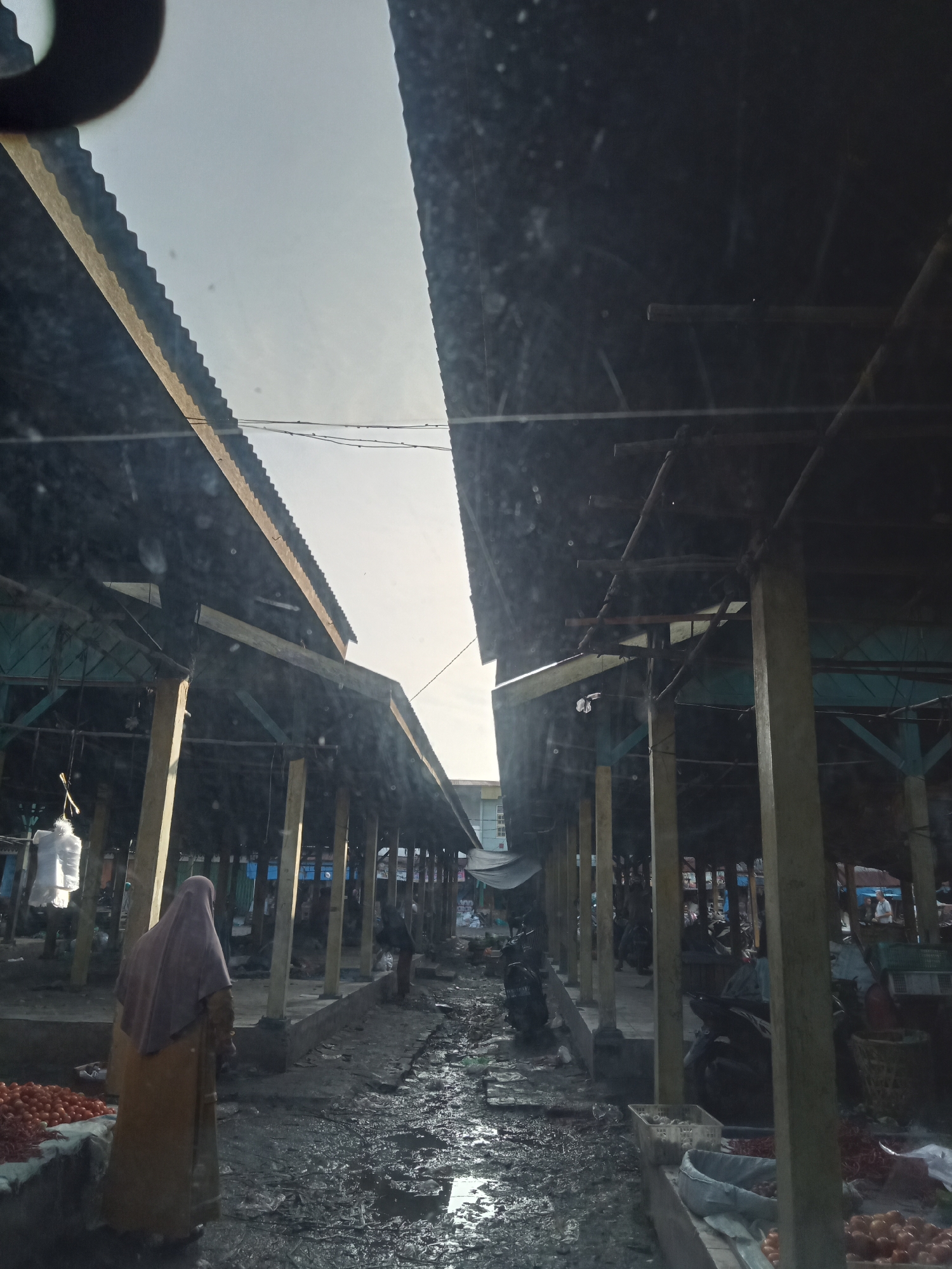 The image depicts an outdoor market scene in Ujung Tanjung, Indonesia. Two covered stalls with corrugated metal roofs, supported by wooden pillars, face each other, creating an aisle. The ground is muddy and uneven, with puddles of water, suggesting recent rain. Stalls are lined with various goods, including piles of tomatoes and other produce. In the left stall, a person wearing a hijab and a yellow garment stands facing away from the camera, near a display of tomatoes. Several motorcycles are parked in the aisle. The overall lighting suggests it is either early morning or late afternoon.