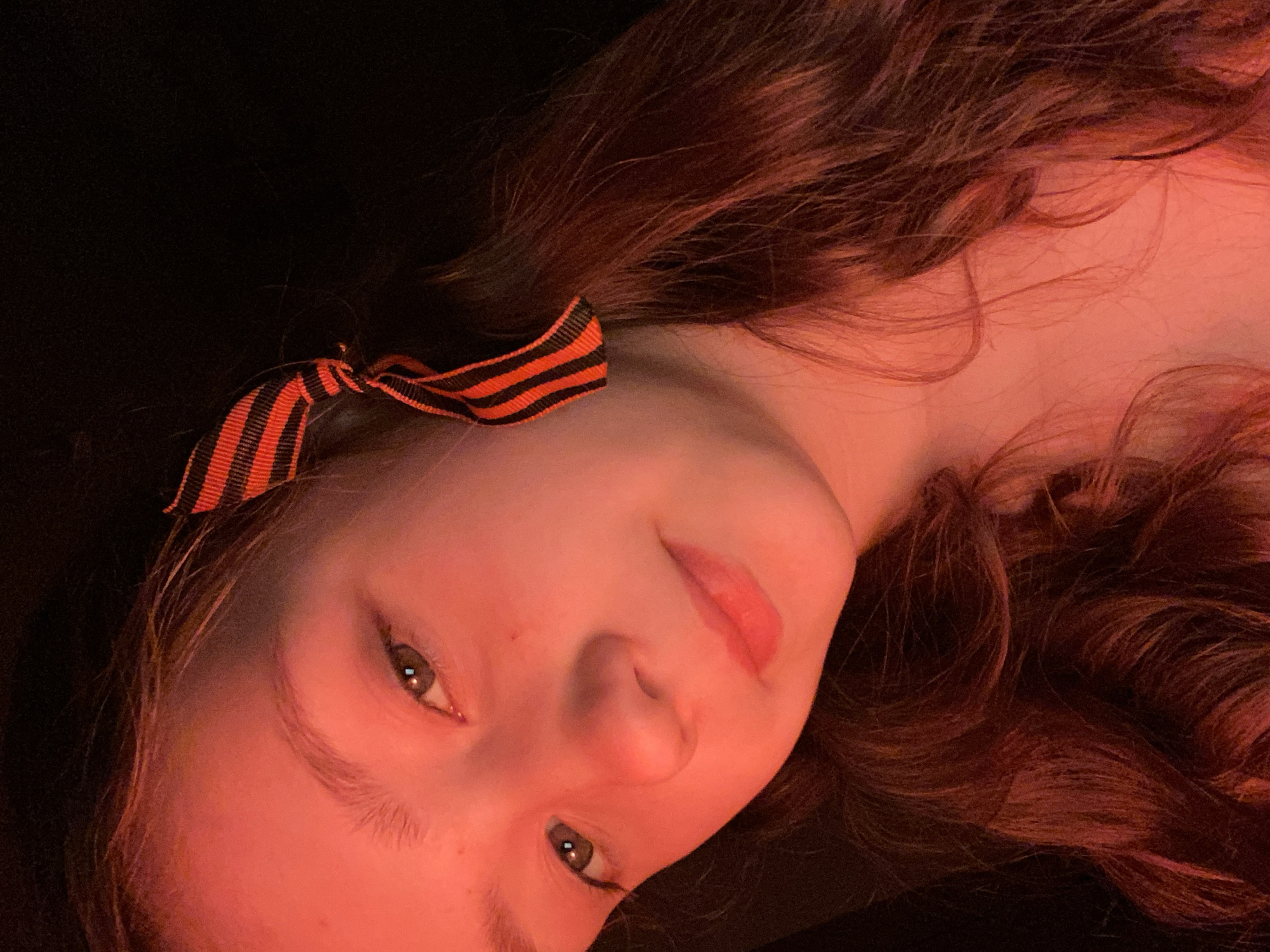 A close-up portrait shows a young woman against a black background. She is lying on her side, with her head angled slightly upward. She has long, wavy brown hair, fair skin, and light-colored eyes. A black and orange striped ribbon is tied in her hair, near her forehead. The lighting casts a reddish hue over the scene. The location is Unknown, Unknown. There are no other people, subjects, objects, actions, or interactions visible.