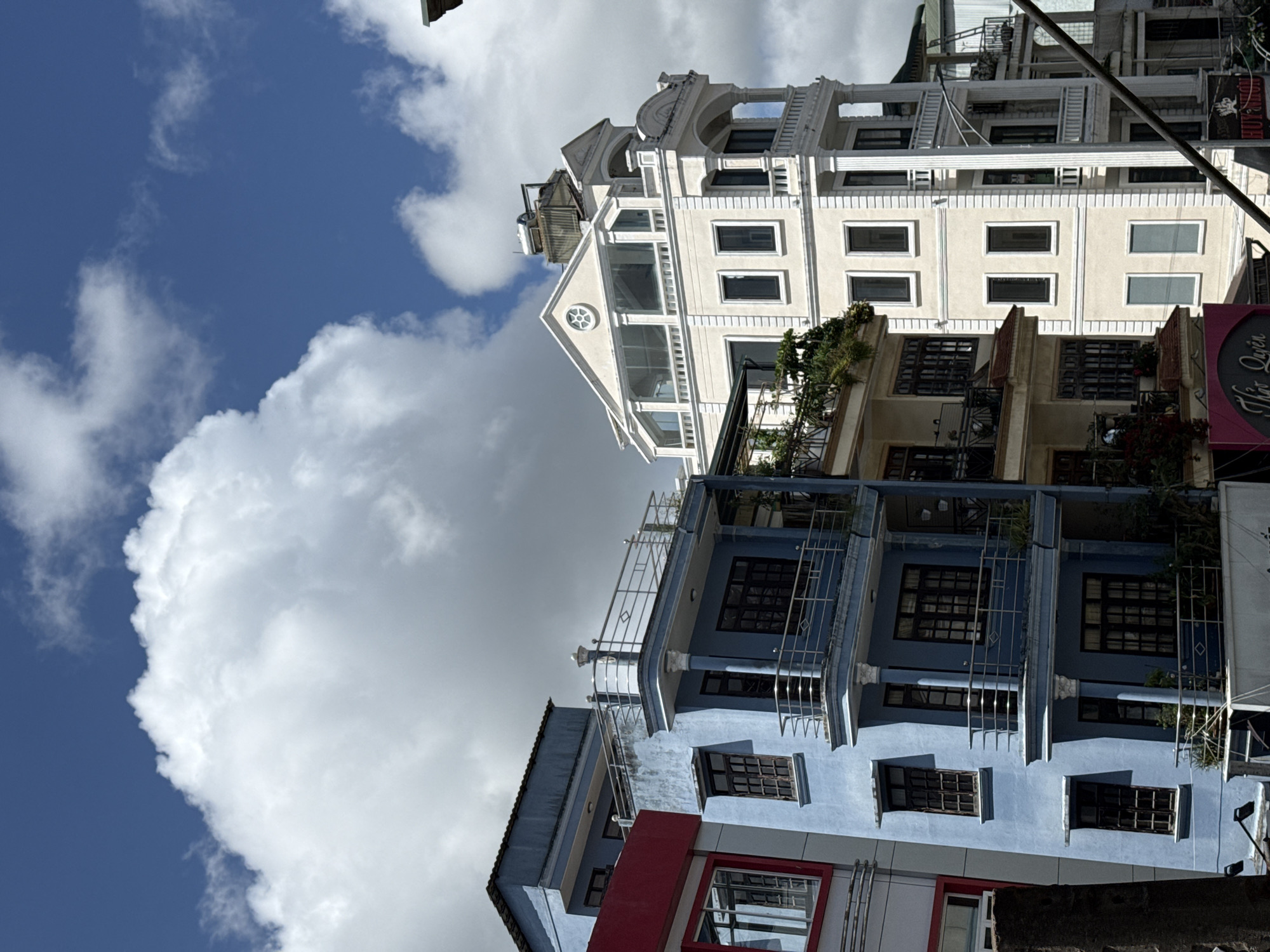 The image shows a street in Da Lat, Vietnam, with several multi-story buildings. The buildings are of various architectural styles and colors, including light blue, cream, and red. Some of the buildings have balconies with metal railings, and others have large windows. The sky is blue with fluffy white clouds. There are no people or vehicles visible in the image. The overall impression is of a quiet, residential street in a city.