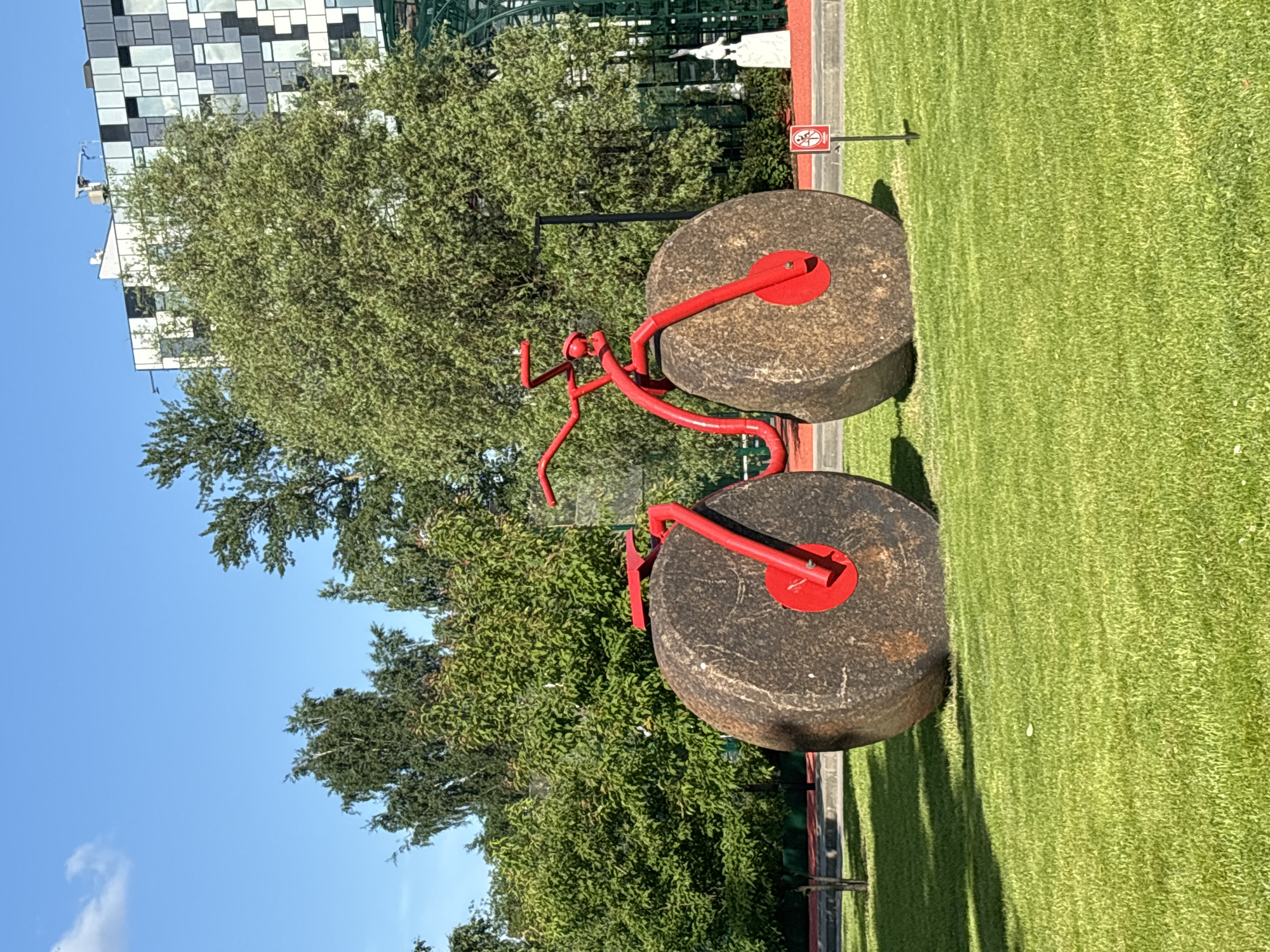 A quirky, abstract sculpture of a bicycle made of red metal and large, round stone wheels sits on a lush green lawn. The bicycle is positioned centrally in the frame, with its front wheel closer to the viewer. Behind the sculpture, a dense line of green trees stretches across the background, partially obscuring a modern building with a distinctive black and white tiled facade. The sky above is a clear, bright blue with a single, wispy white cloud. To the right of the sculpture, a small red sign with a white pictogram is planted in the grass. In the far background, a white statue of a bird or animal on a pedestal is visible. The image captures a sunny day in a park-like setting. This sculpture is located in front of the Dynamo Sports Palace in St. Petersburg, Russia.